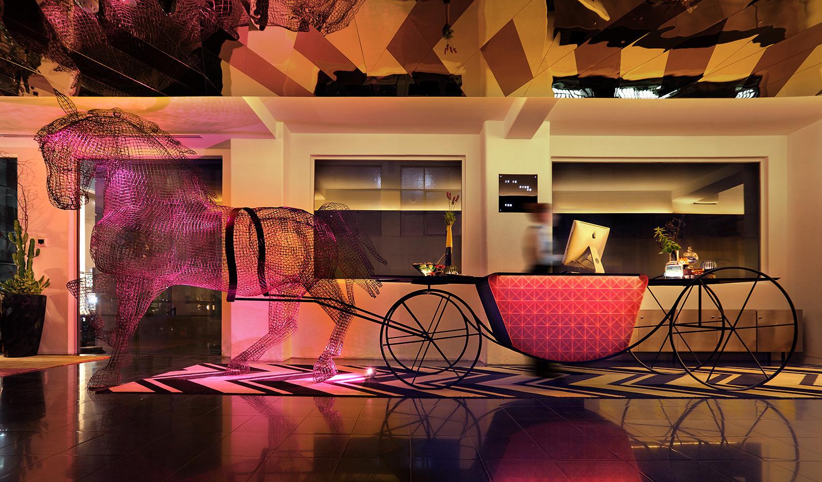 A hotel lobby at night with a wire sculpture of a horse and carriage used as a reception desk, lit in pink and gold, with a staff member blurred in motion behind it.