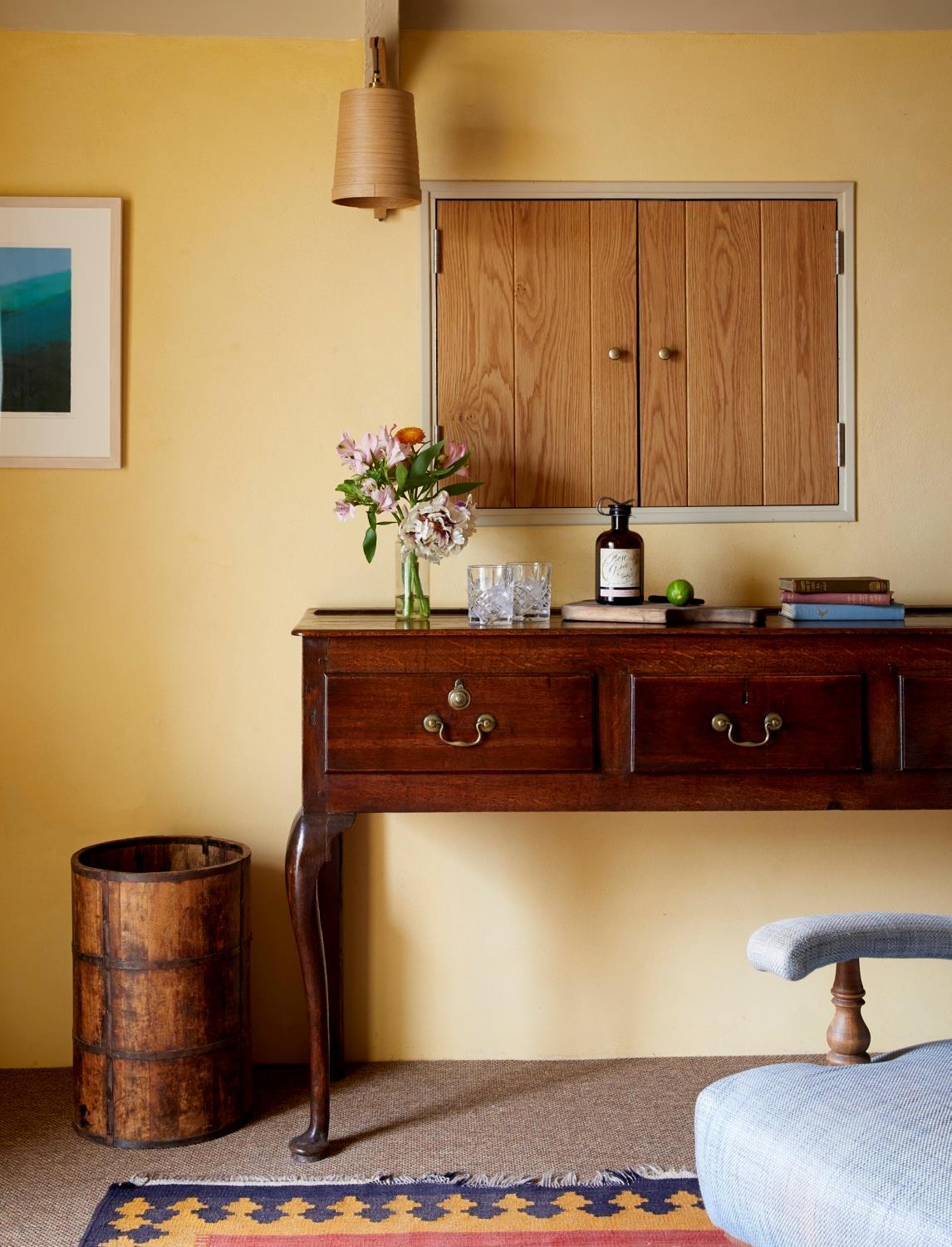 A wooden chest of drawers with flowers in a jug under a painted cupboard, in a room with pale yellow walls.