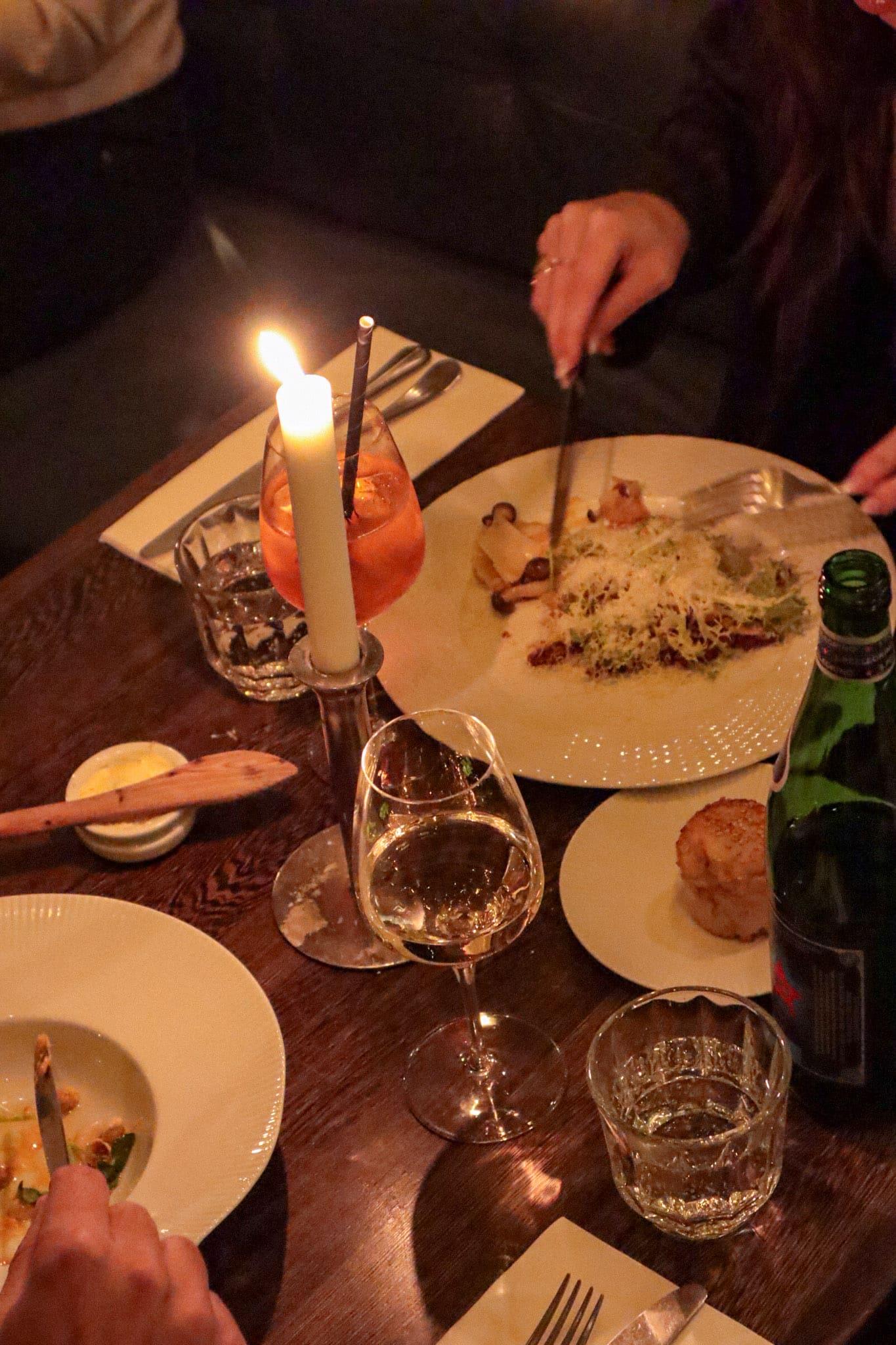 A candlelit restaurant table set with wine glasses, small plates of food and a sparkling water bottle, with two people's hands reaching across it.