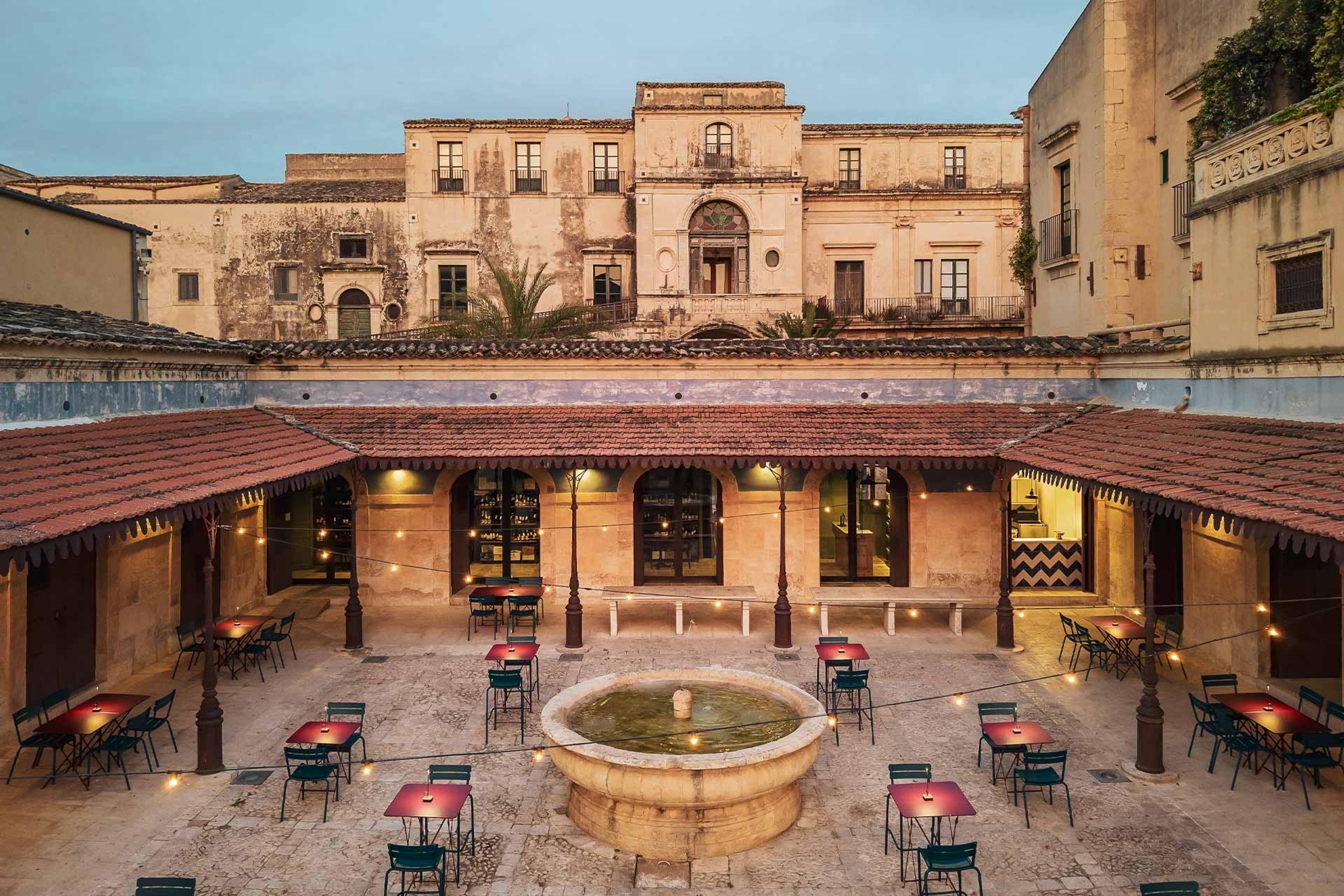 An elevated view of a stone courtyard with a round fountain and red cafe tables, string lights strung beneath a weathered baroque palazzo at dusk.