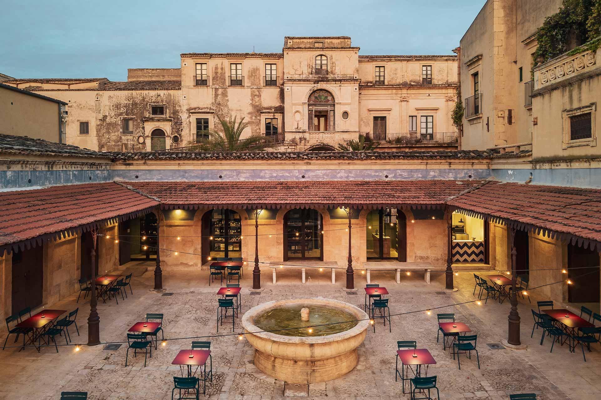 An elevated view of a stone courtyard with a round fountain and red cafe tables, string lights strung beneath a weathered baroque palazzo at dusk.