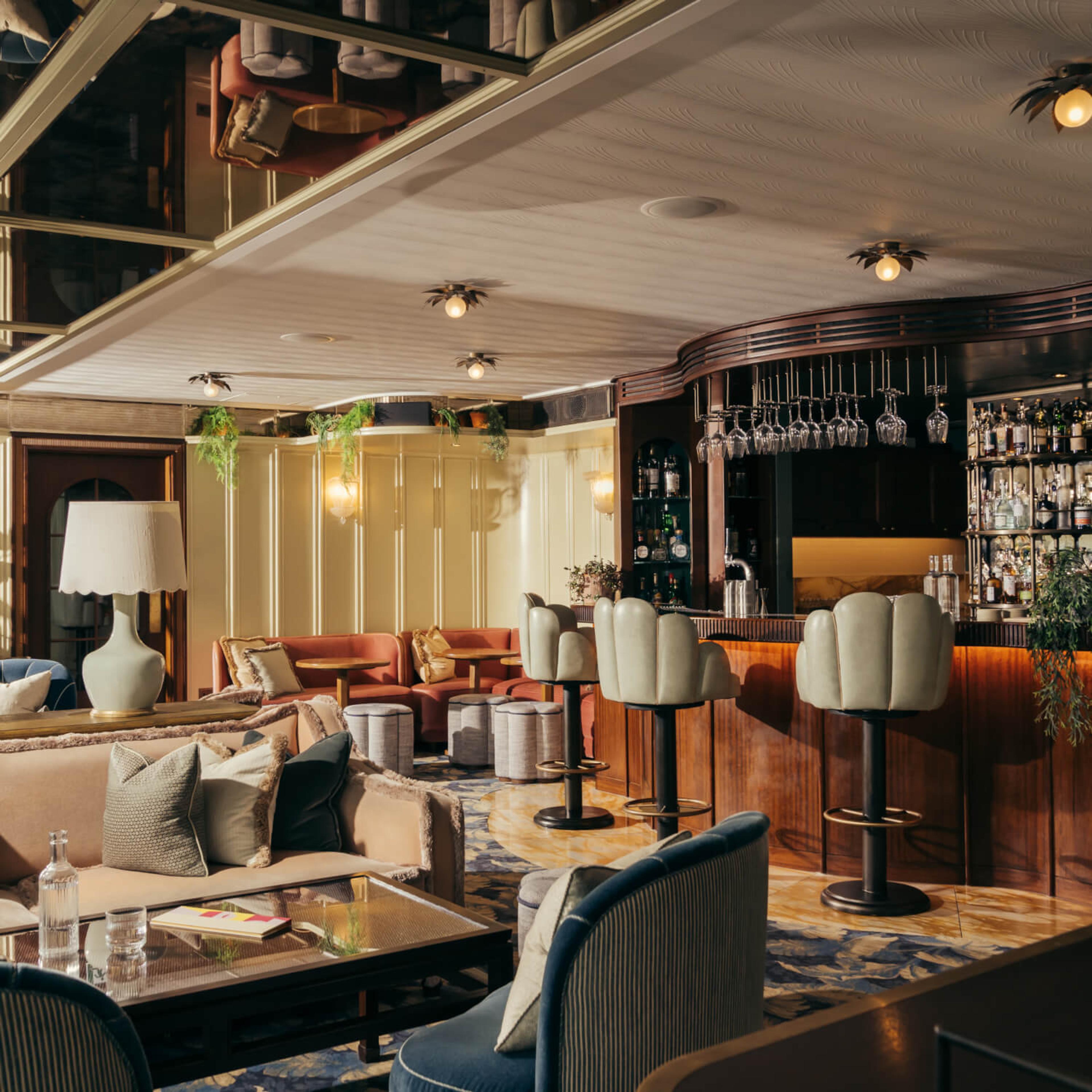 a hotel bar with a curved wooden counter, hanging glassware, scalloped stools and low lounge seating under warm lighting