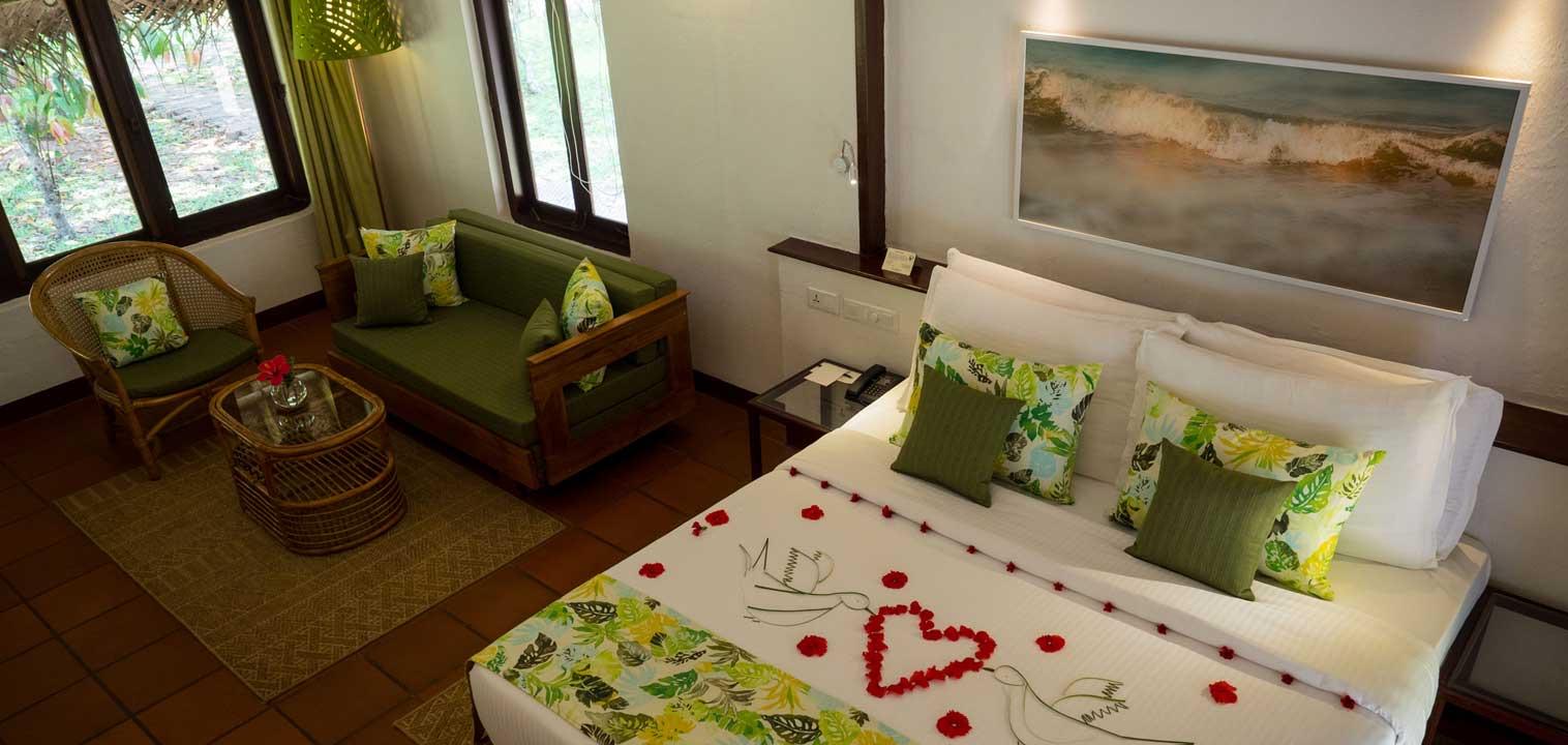 A hotel room in green tones with a sofa, armchair and a bed dressed with a heart made of rose petals, seen from above.