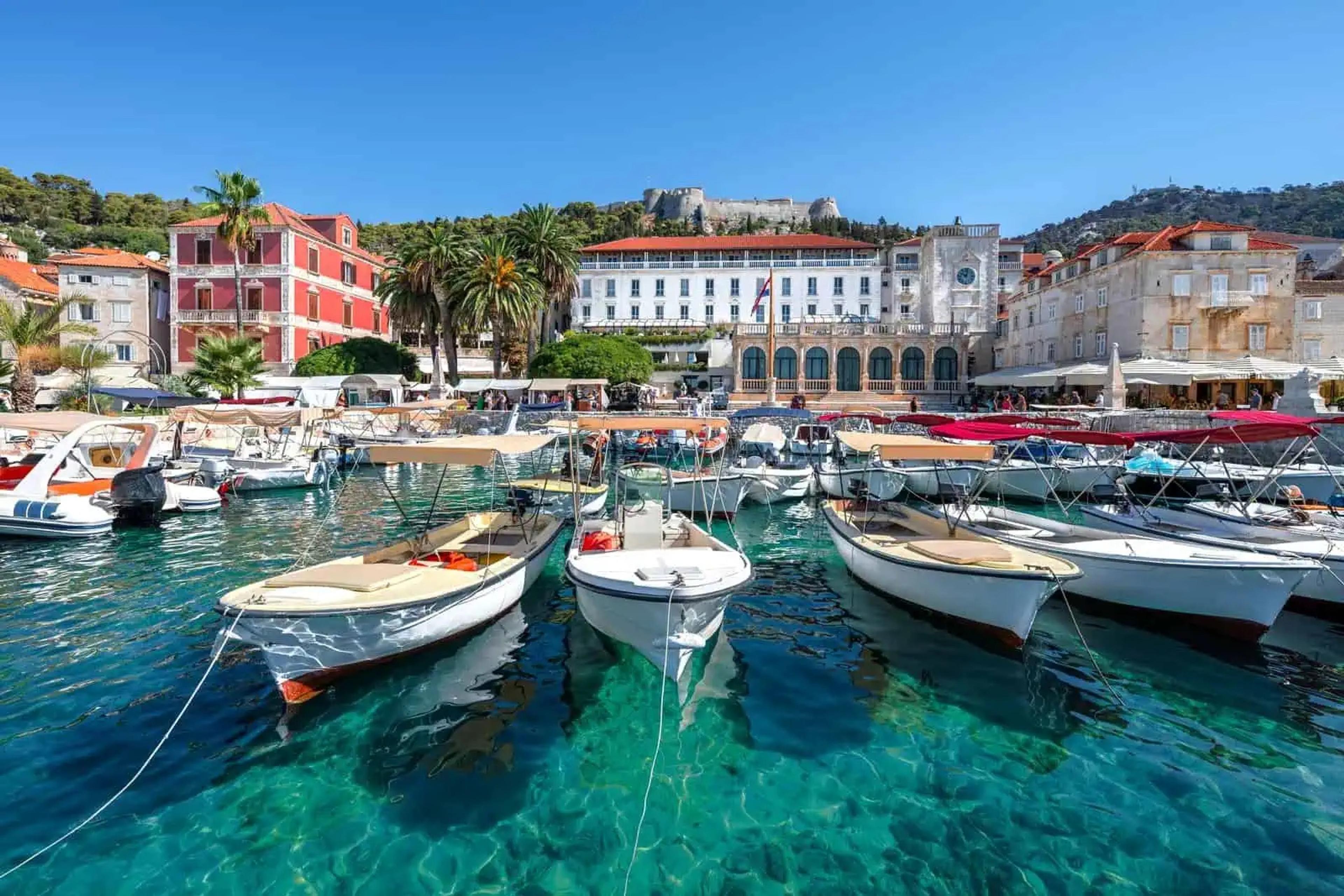 Small boats moored in clear turquoise water along the Riva in Hvar Town, the fortress on the hill above