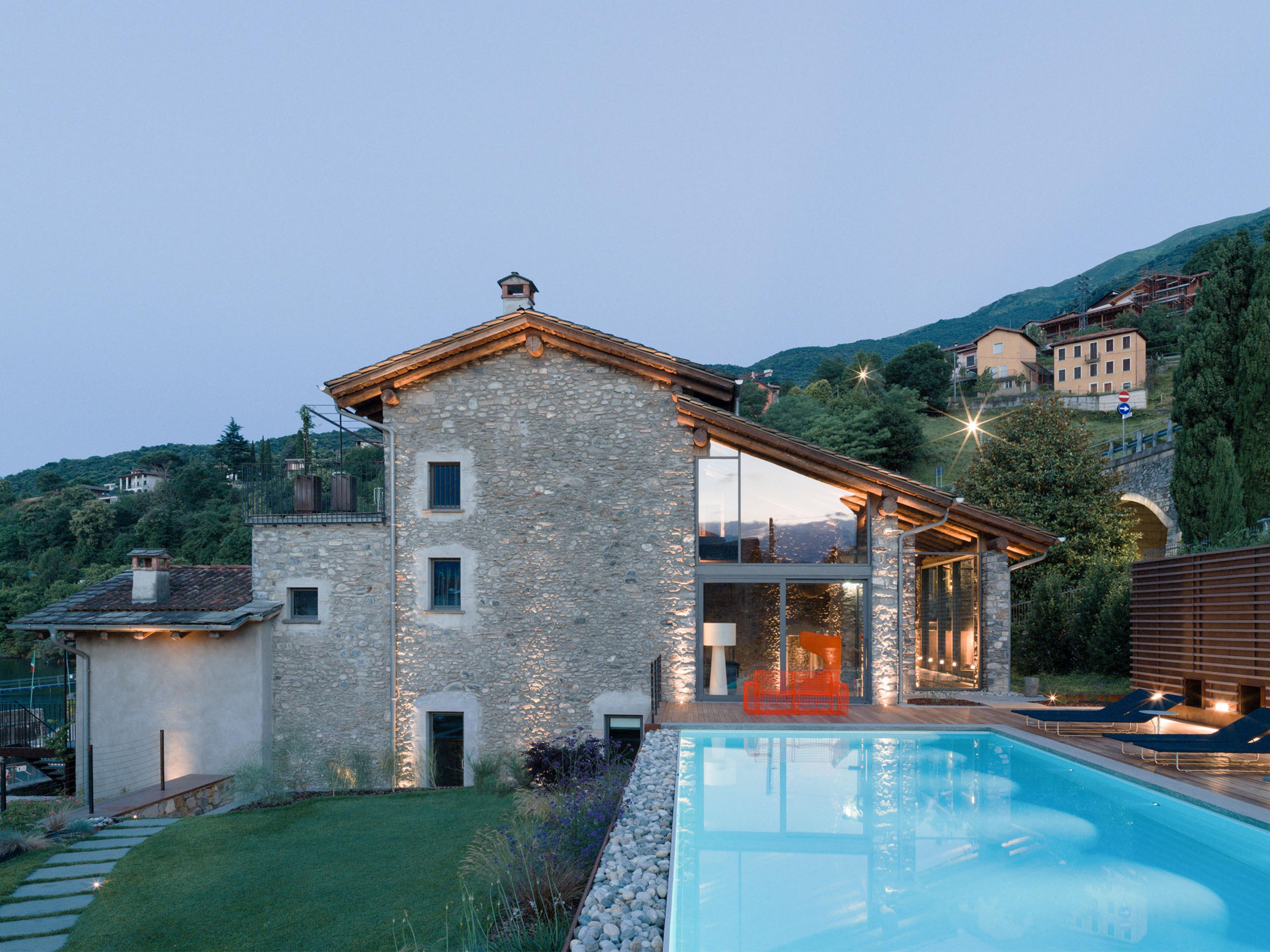 a stone building beside a lit swimming pool at dusk, with an orange chair on the deck and the hillside and village lights behind it