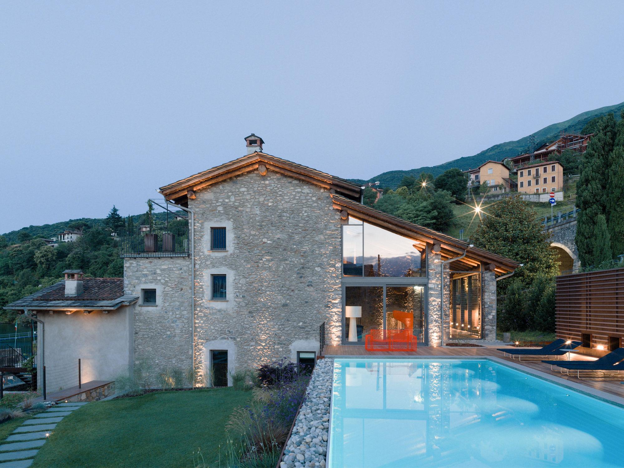 a stone building beside a lit swimming pool at dusk, with an orange chair on the deck and the hillside and village lights behind it