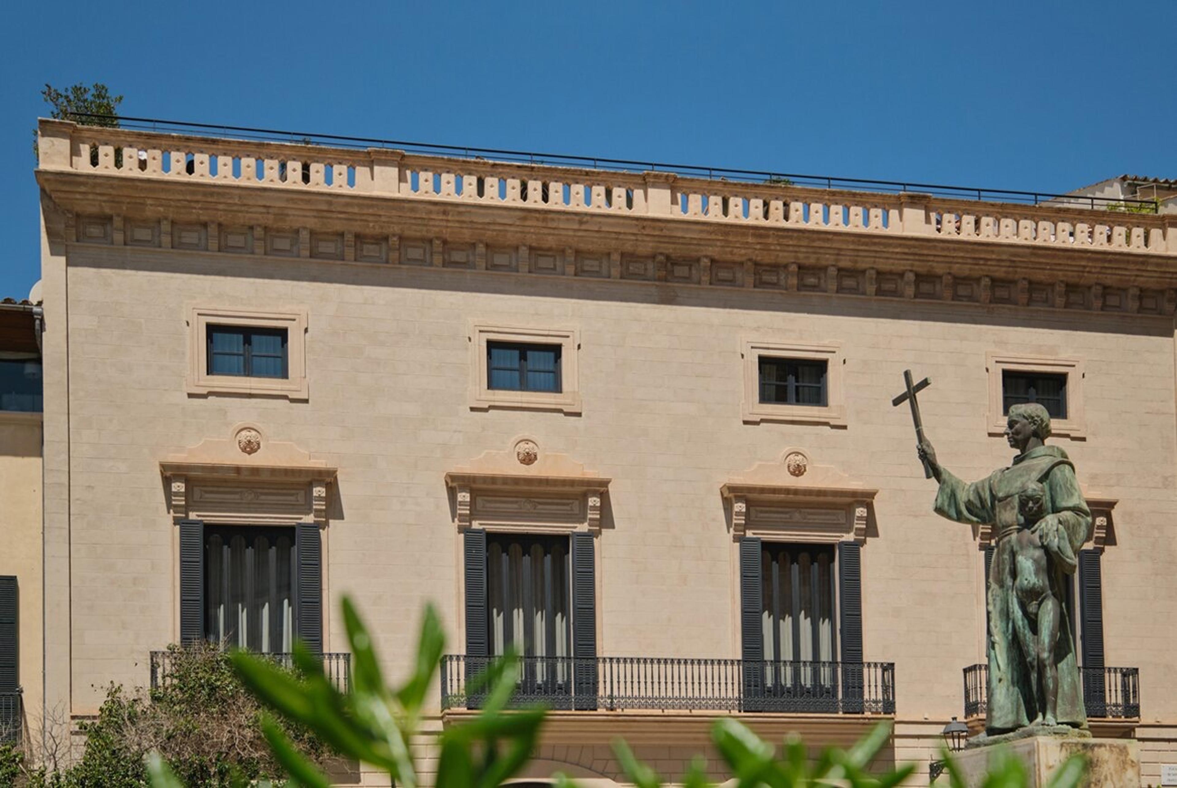 A cream stone building with shuttered windows and wrought iron balconies, seen past a bronze statue of a robed figure holding a cross, with leaves crossing the foreground.