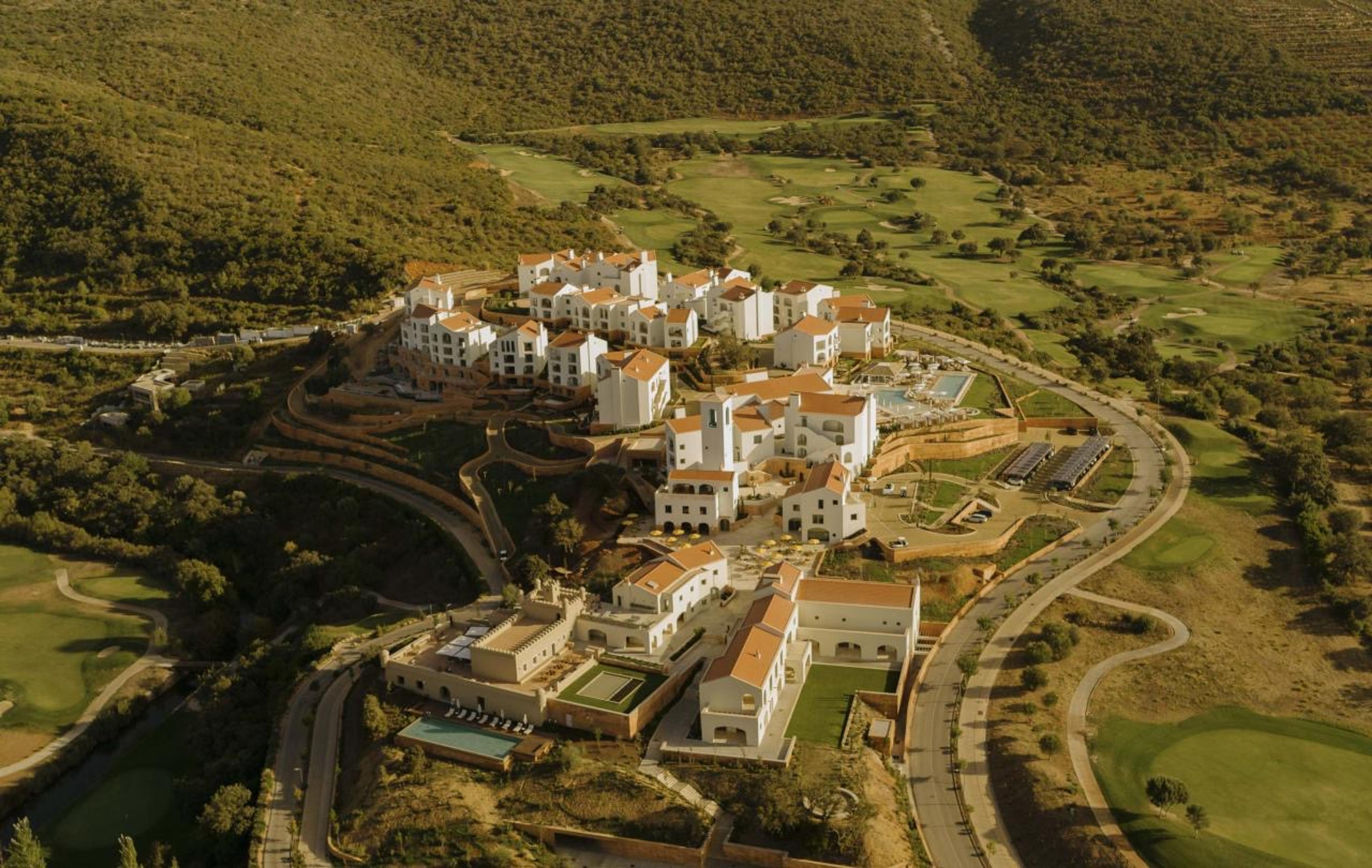 An aerial view of whitewashed, terracotta-roofed resort buildings set in a wooded valley next to a golf course.