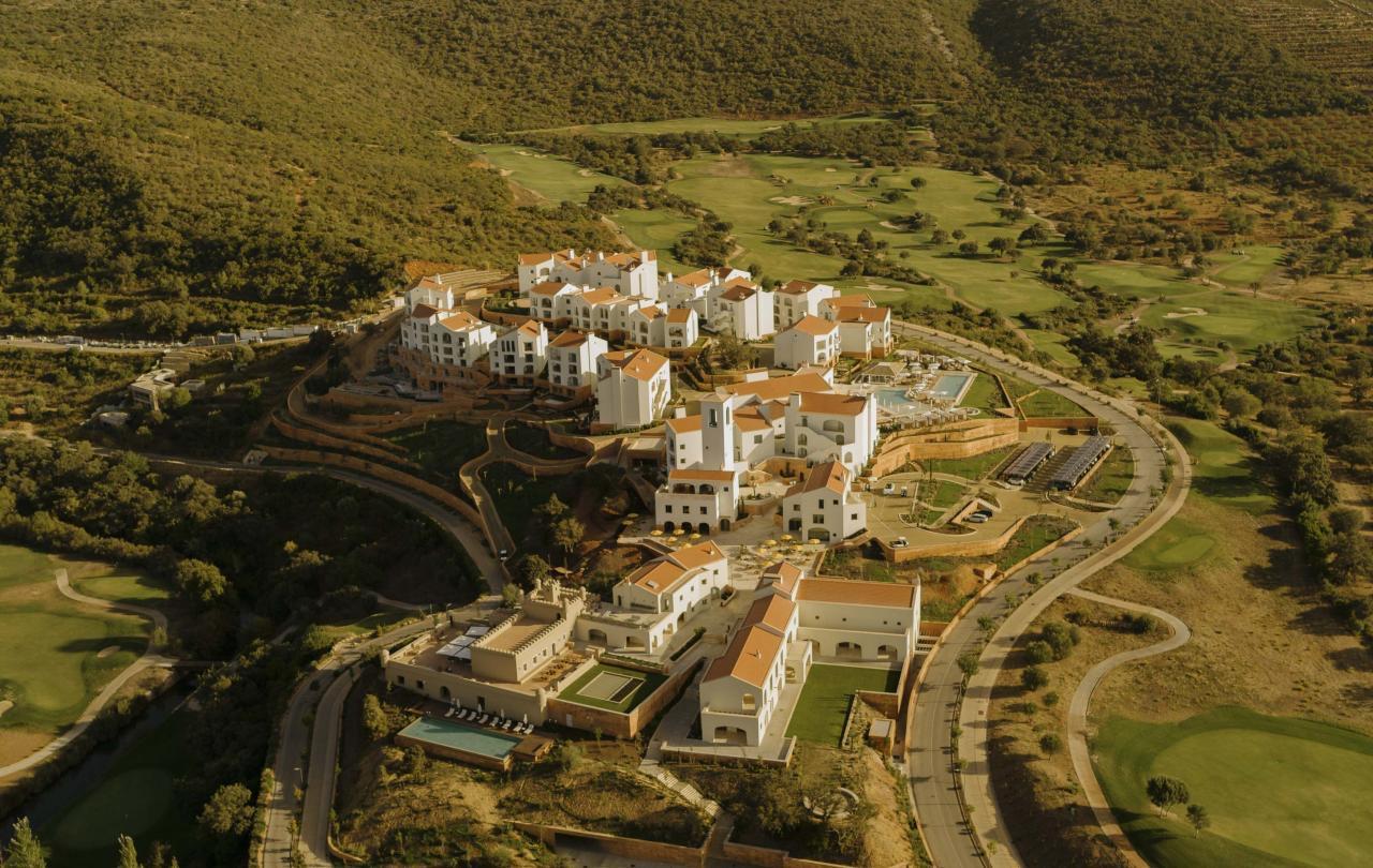 An aerial view of whitewashed, terracotta-roofed resort buildings set in a wooded valley next to a golf course.
