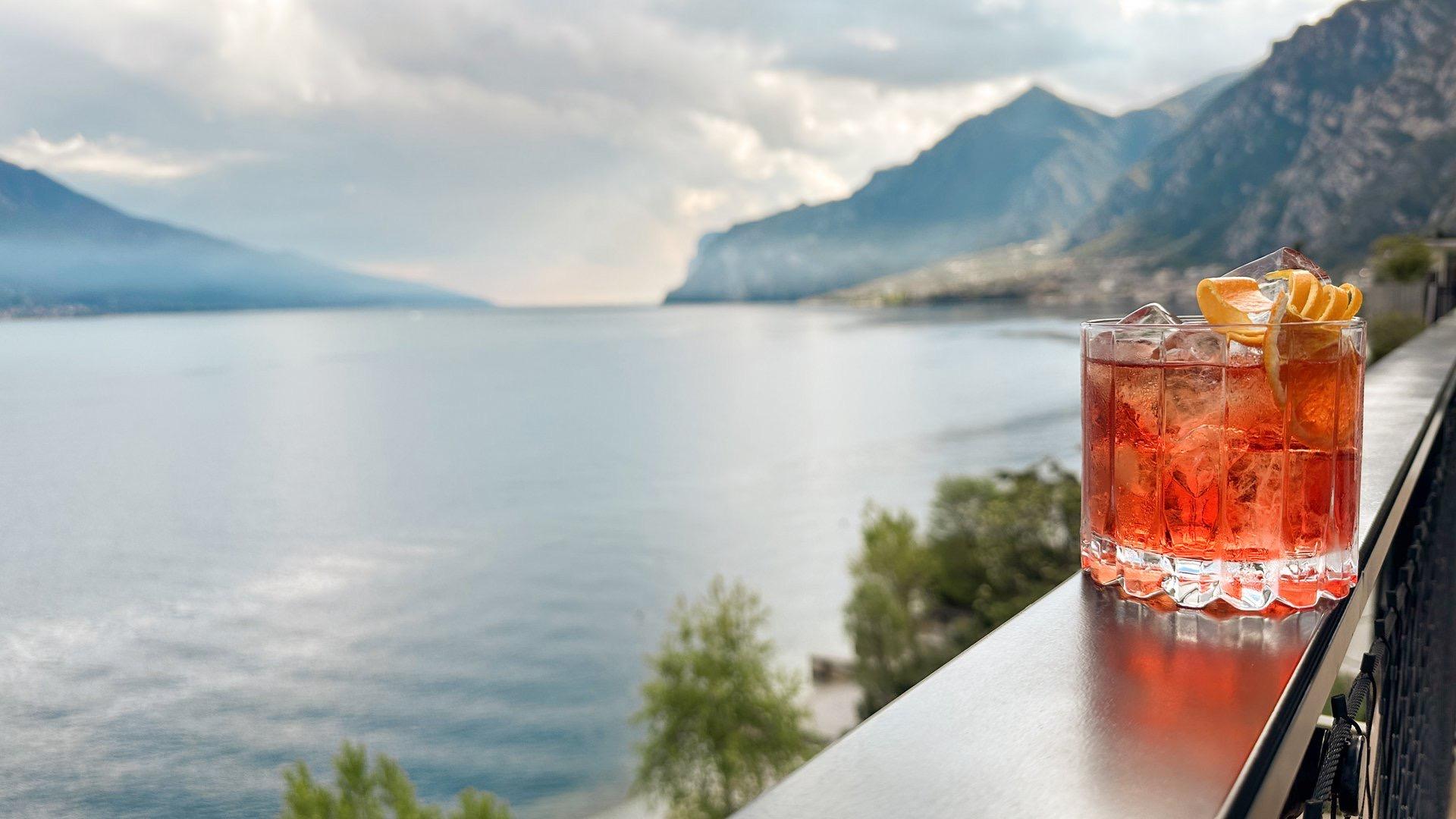 a red cocktail garnished with orange peel standing on a railing above a lake with mountains behind