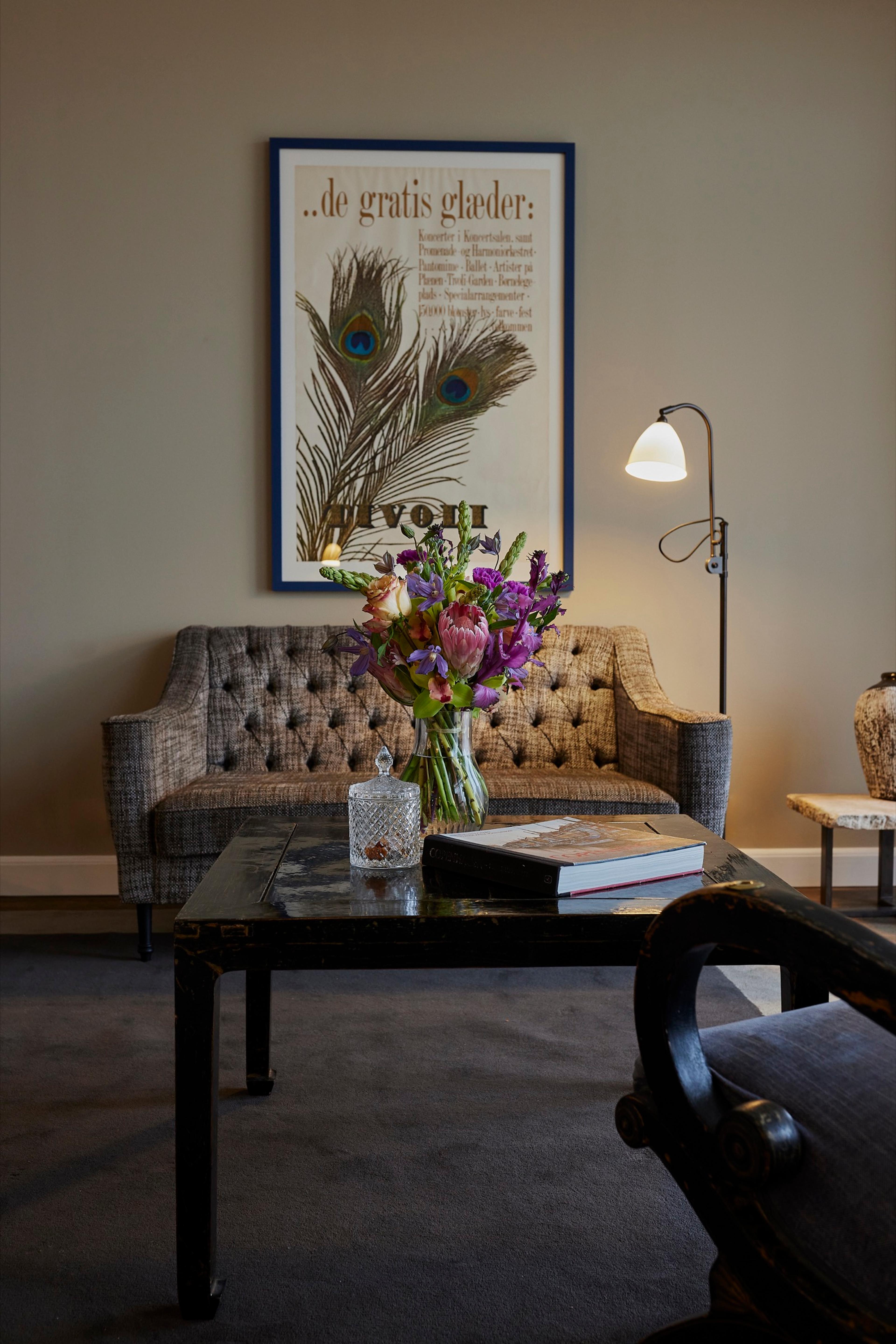 A tufted sofa and a floor lamp sit beside a low table holding a crystal box, a book and a vase of orchids, under a framed vintage Tivoli poster of peacock feathers.