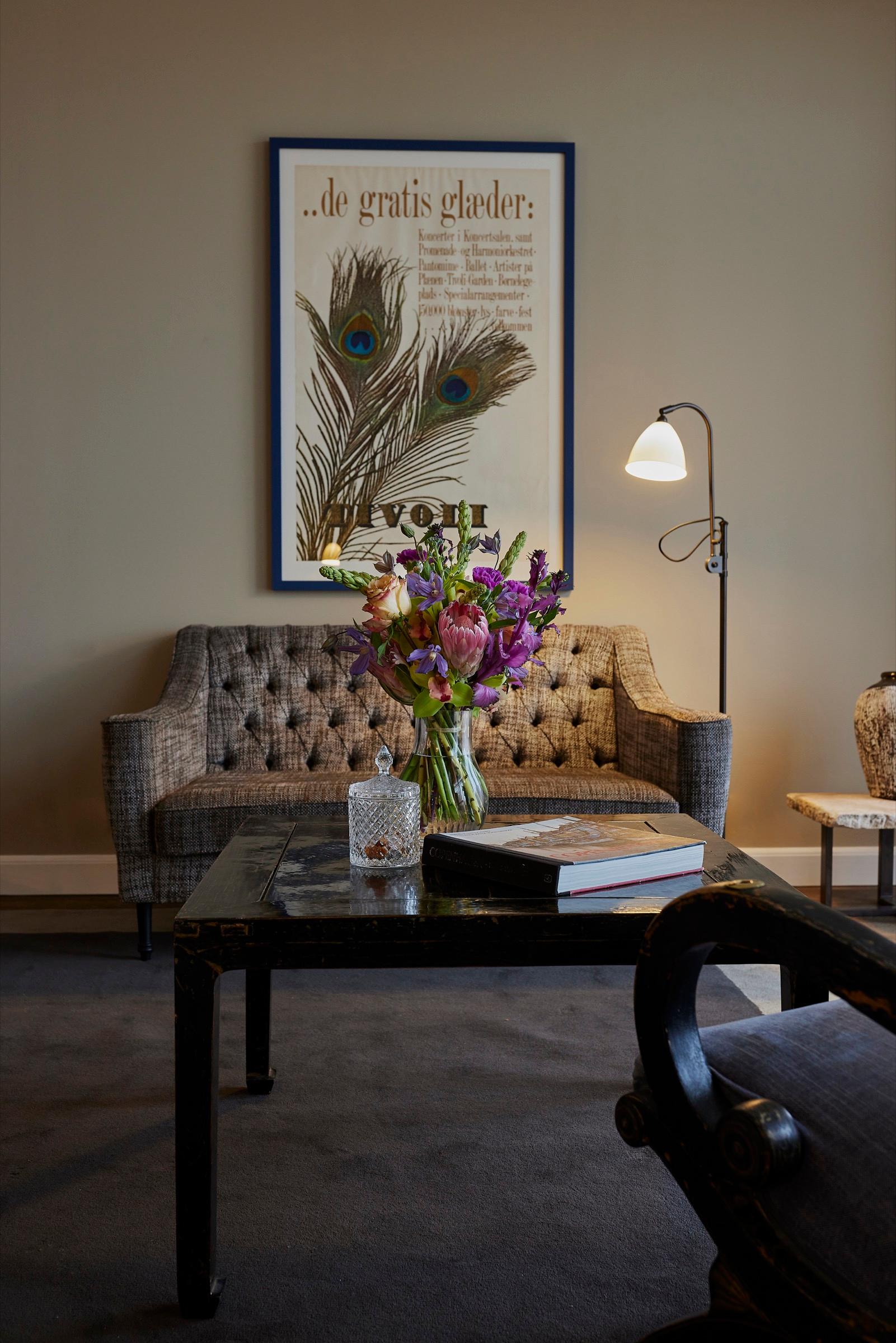 A tufted sofa and a floor lamp sit beside a low table holding a crystal box, a book and a vase of orchids, under a framed vintage Tivoli poster of peacock feathers.