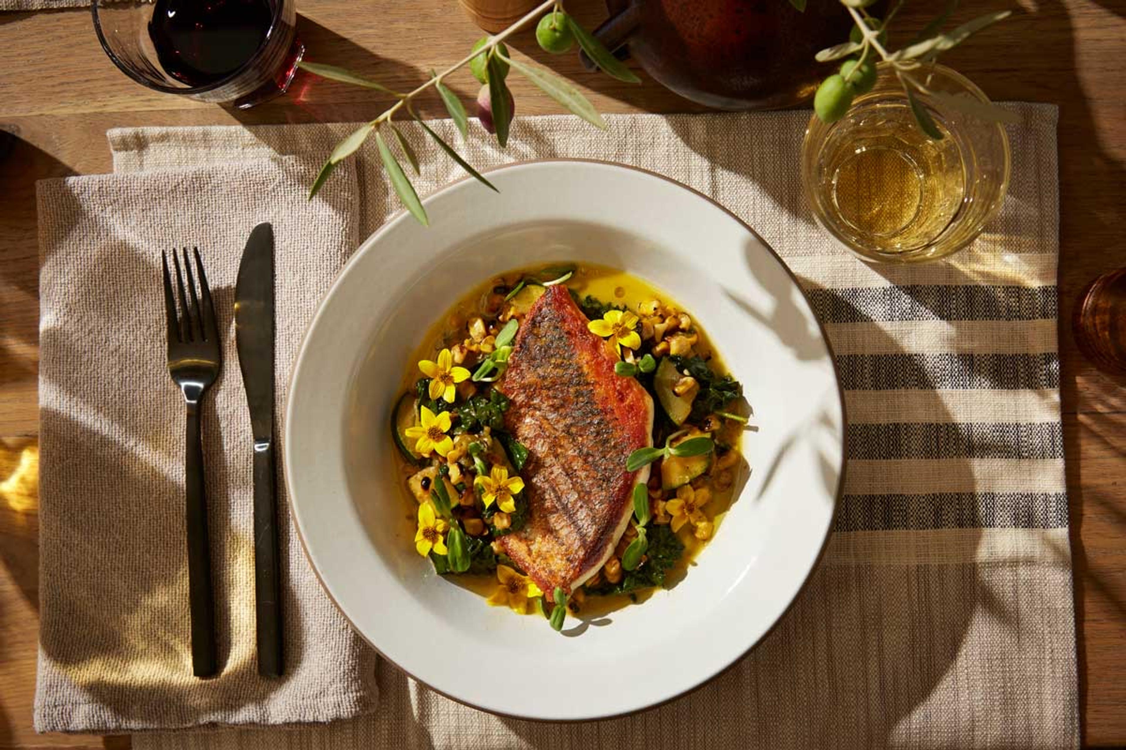 A seared fish fillet with crisp skin on a white plate, with greens and yellow flowers, on a wooden table set with linen napkins and two glasses of wine.