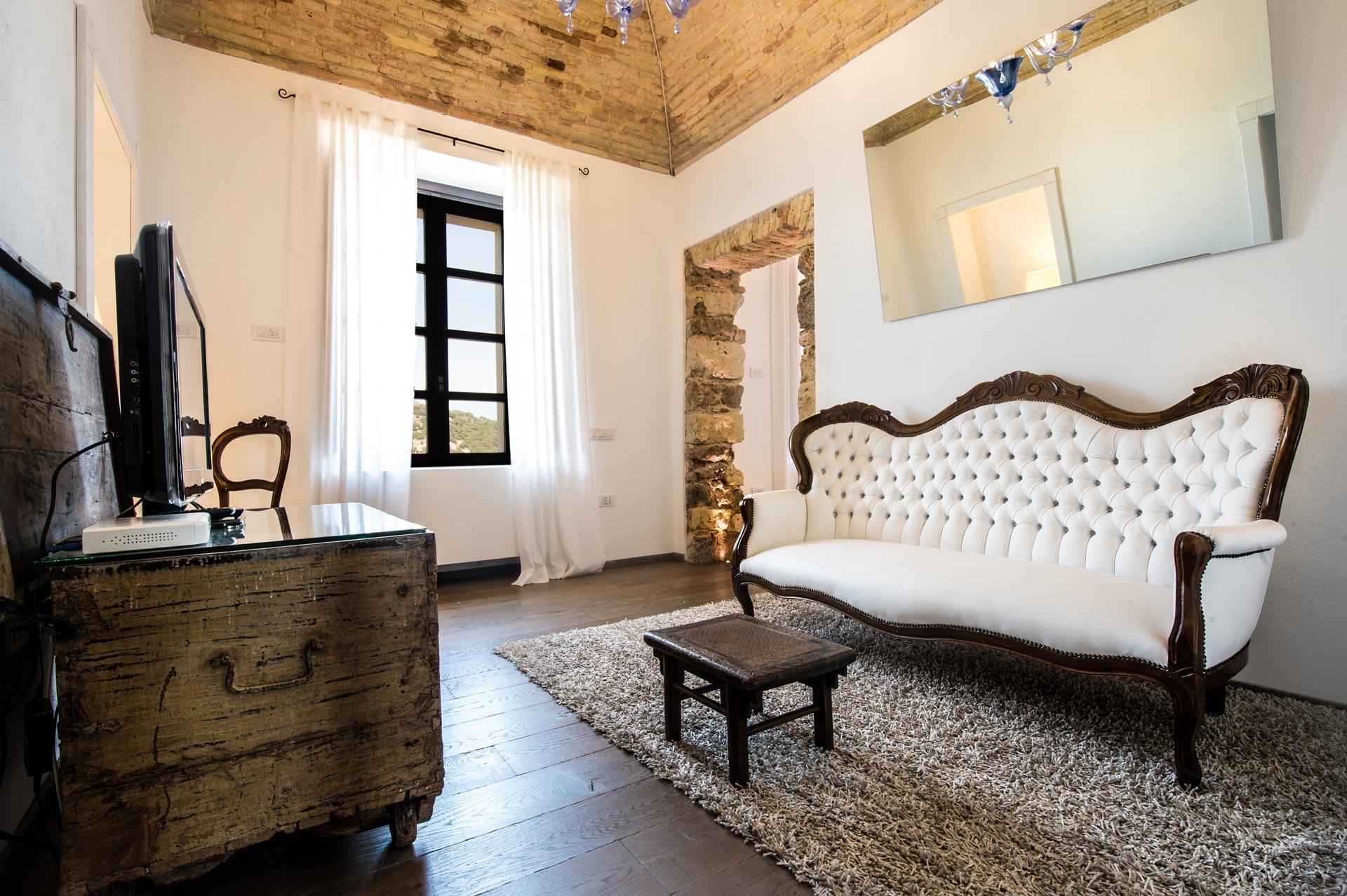 a sitting room with a white tufted antique sofa, a wooden chest with a television, a stone archway and a window with white curtains looking onto greenery