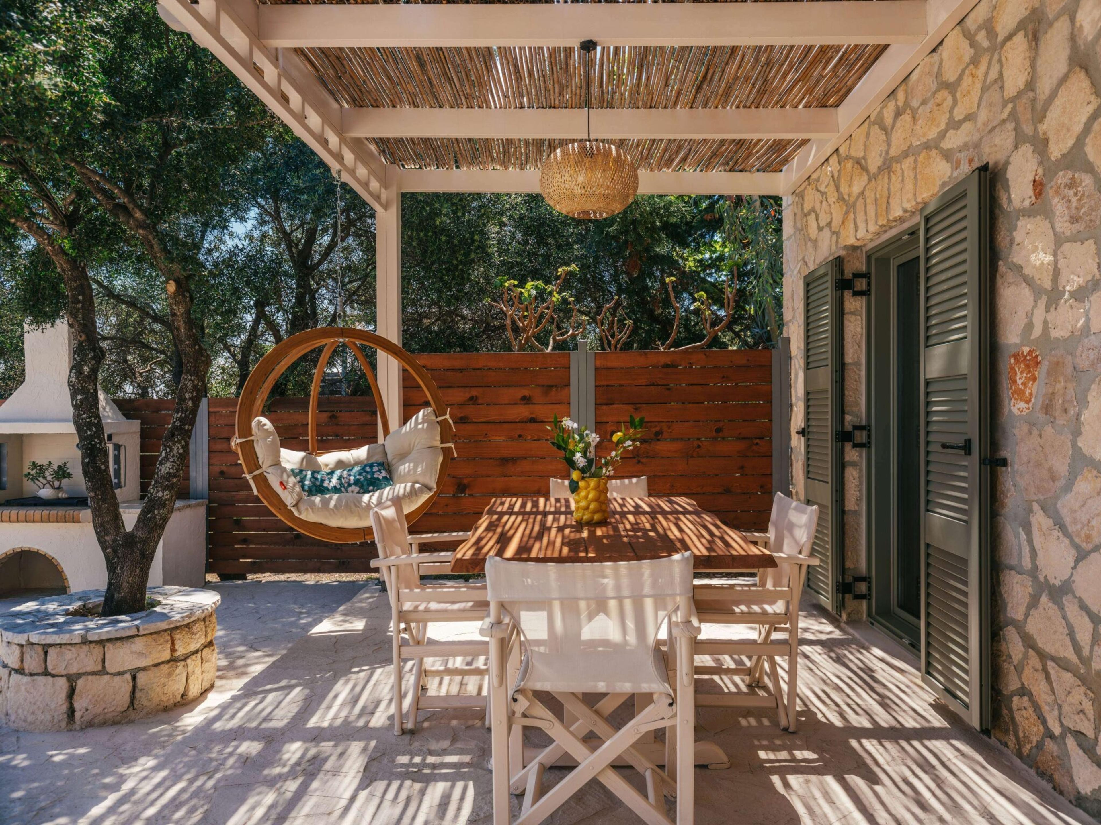 a wooden table laid with white chairs under a reed-covered pergola, a vase of lemons on the table and a stone farmhouse behind