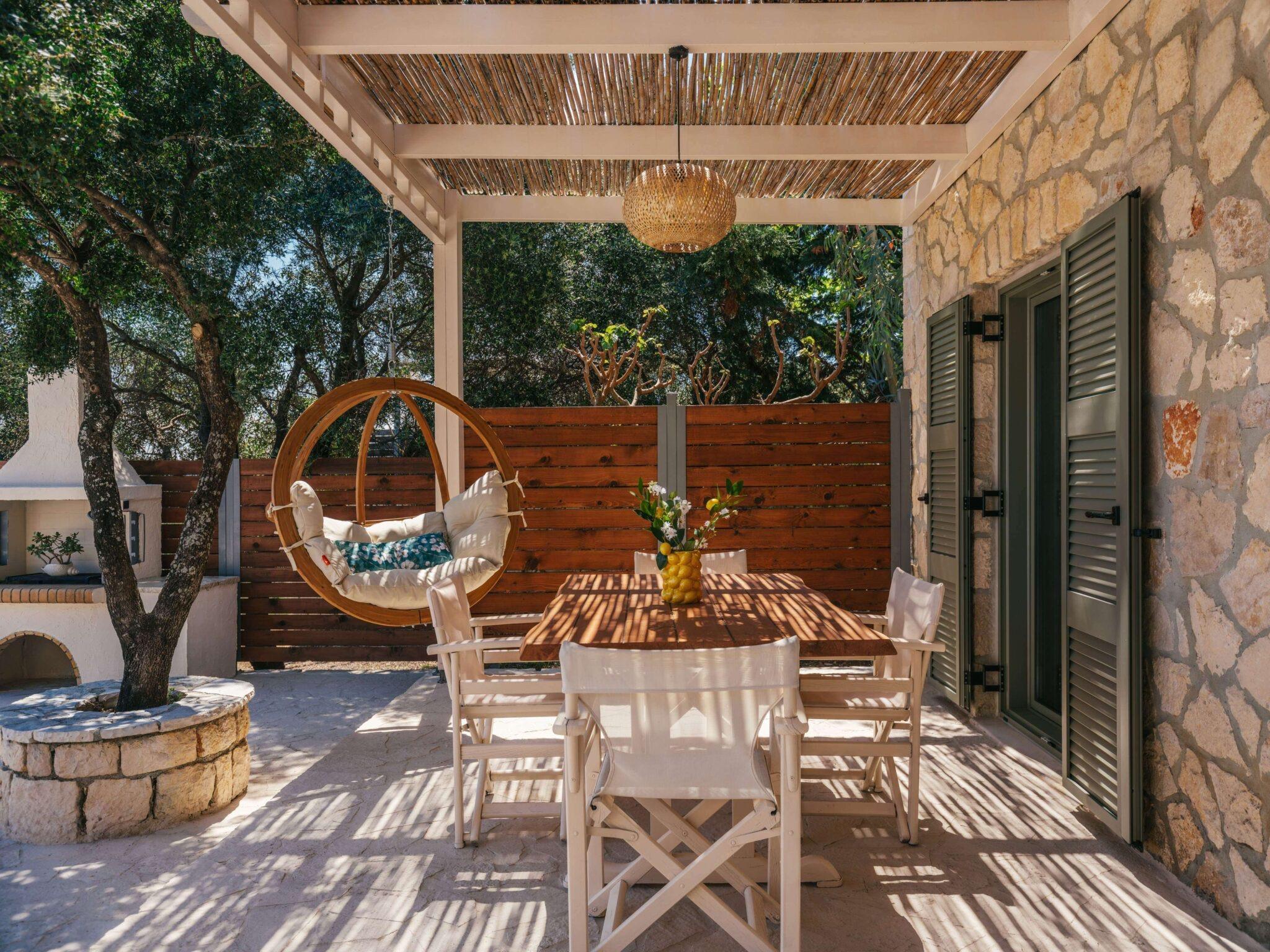 a wooden table laid with white chairs under a reed-covered pergola, a vase of lemons on the table and a stone farmhouse behind
