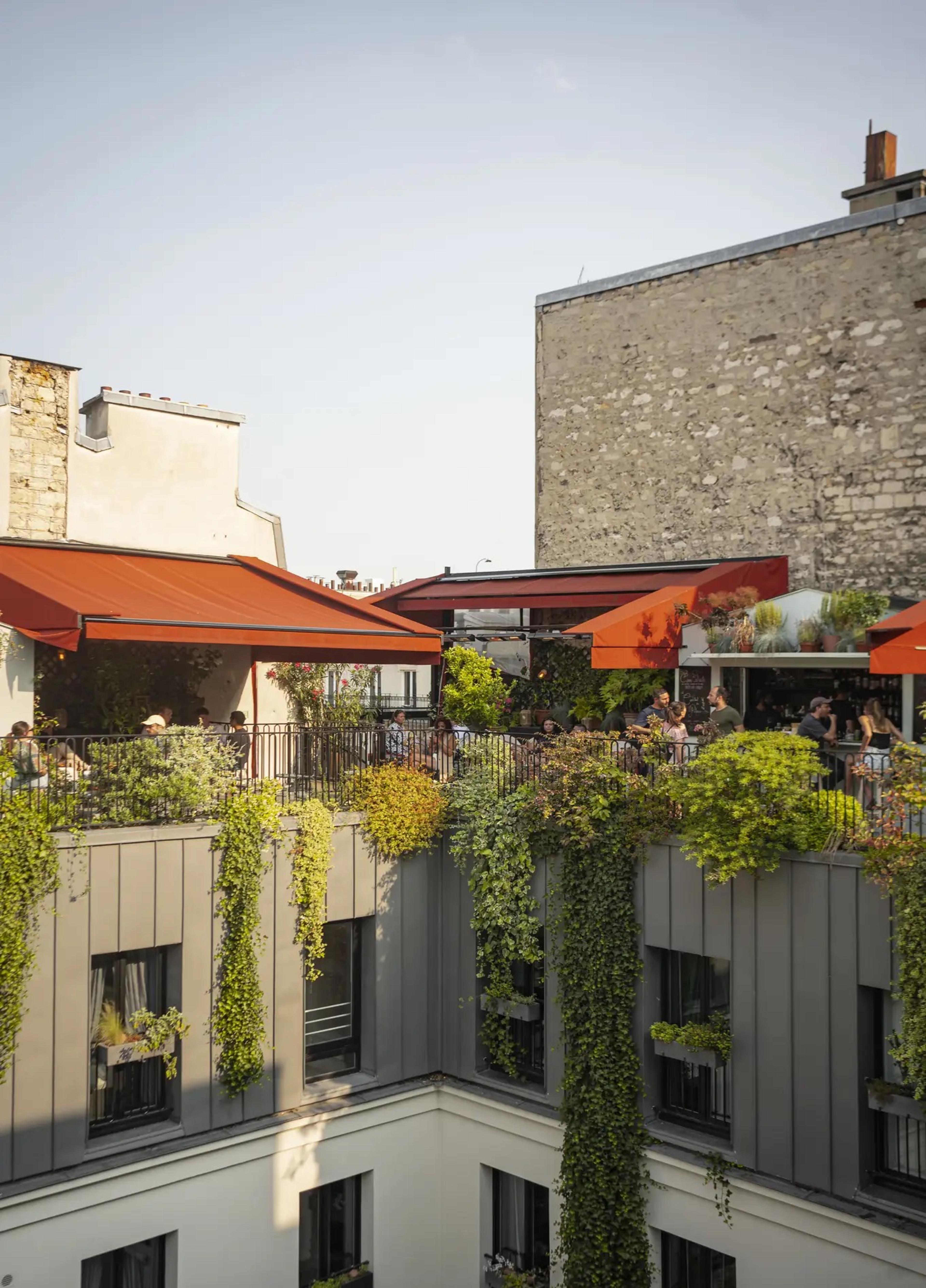 A rooftop terrace with orange awnings and trailing plants sits above Pigalle's rooftops, with people dining at tables in late-afternoon light.