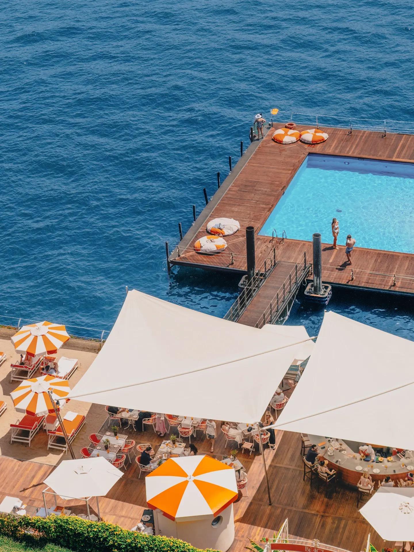 A swimming pool built on a deck over the lake is seen from above, with sun loungers, orange and white umbrellas and a waterside restaurant beside it.