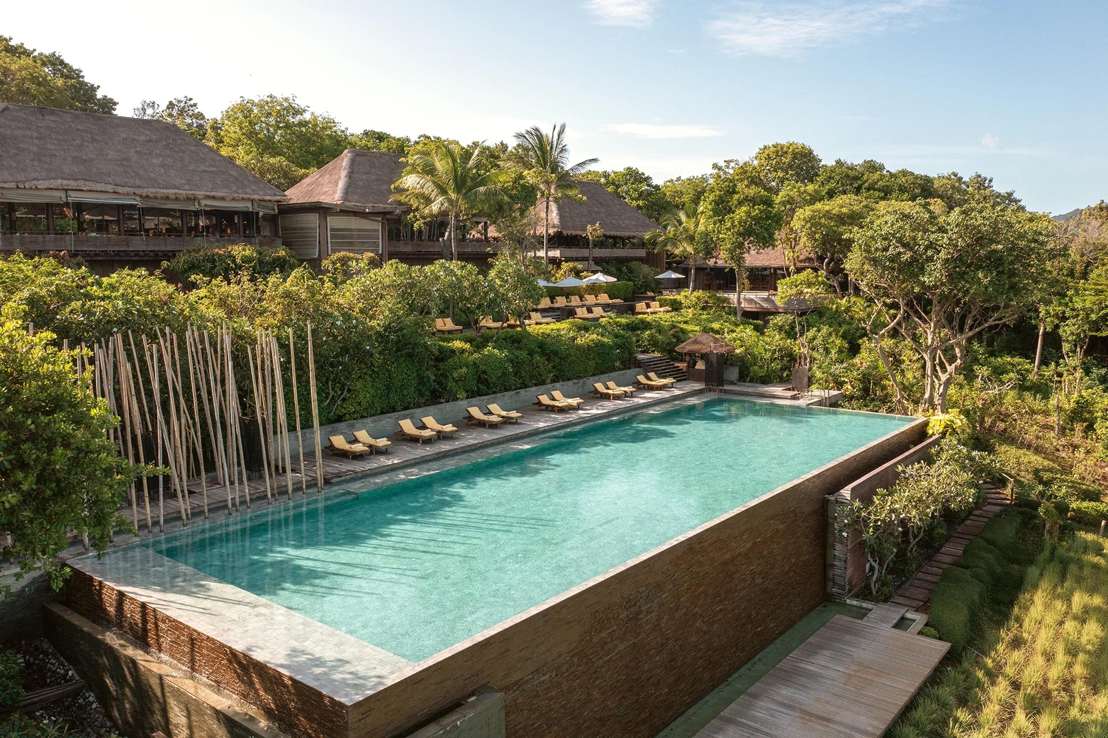An aerial view of a long rectangular pool lined with sun loungers, set among trees in front of a row of thatched resort buildings.