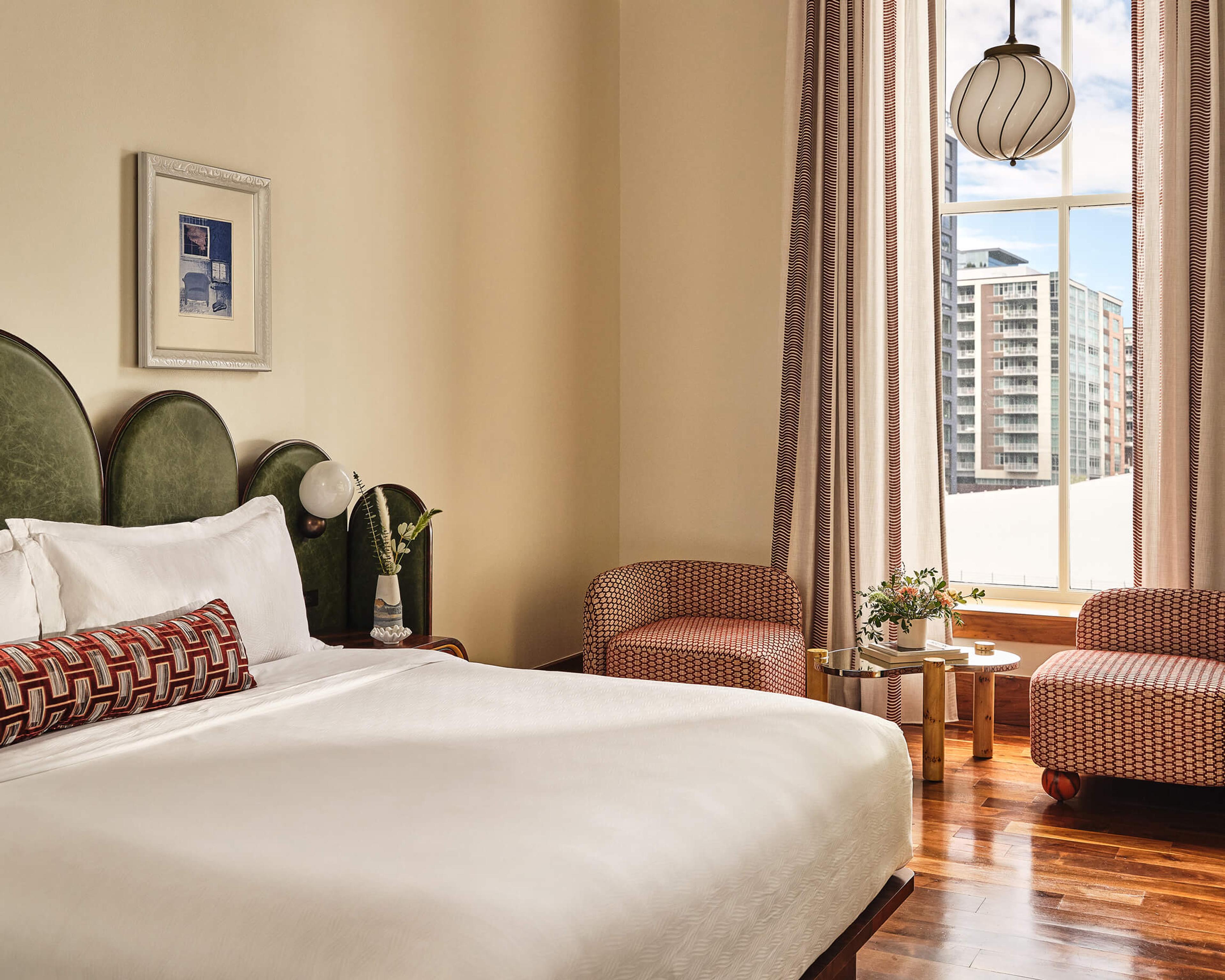 A bed with a green arched headboard beside a tall window, with two patterned armchairs and a view of tall city buildings through the glass.