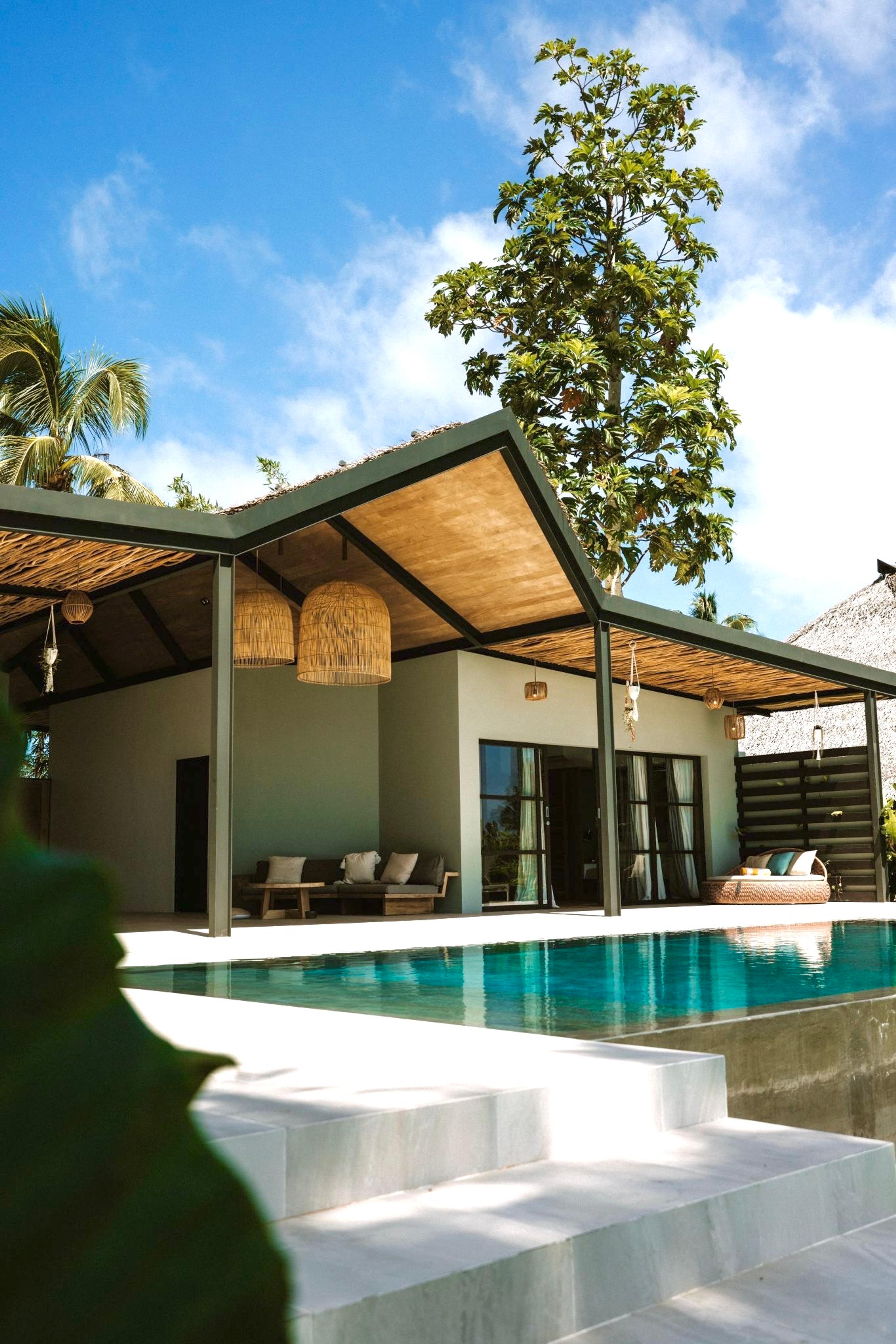 A villa with a dark pergola roof over its terrace, seen past a tree beside its pool under a blue sky.