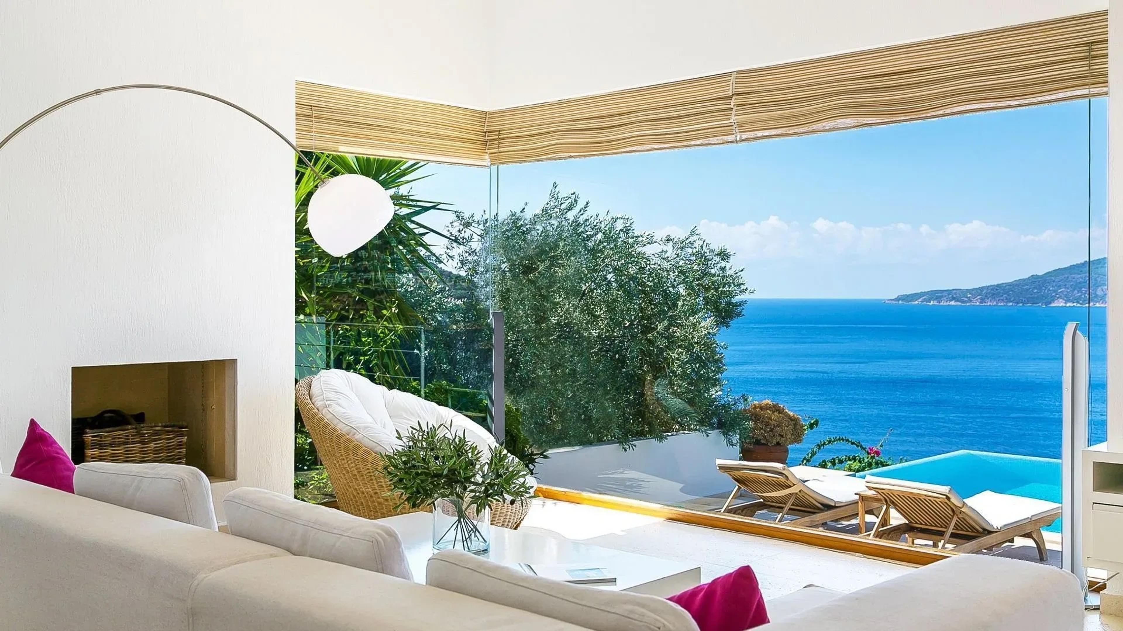 A white living room with a cream sofa, pink cushions and a fireplace, its glass wall looking out to a pool and the sea.