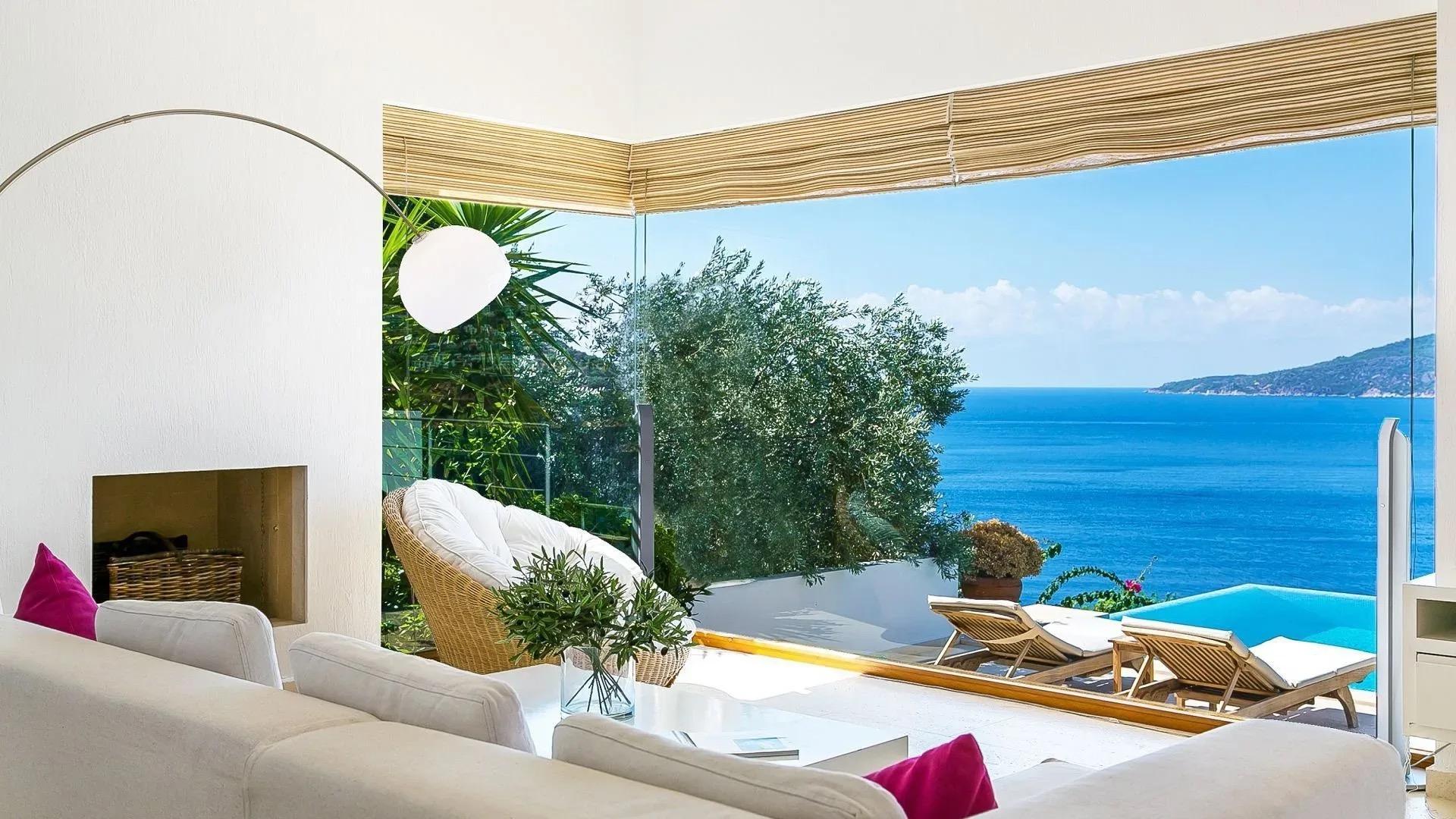 A white living room with a cream sofa, pink cushions and a fireplace, its glass wall looking out to a pool and the sea.