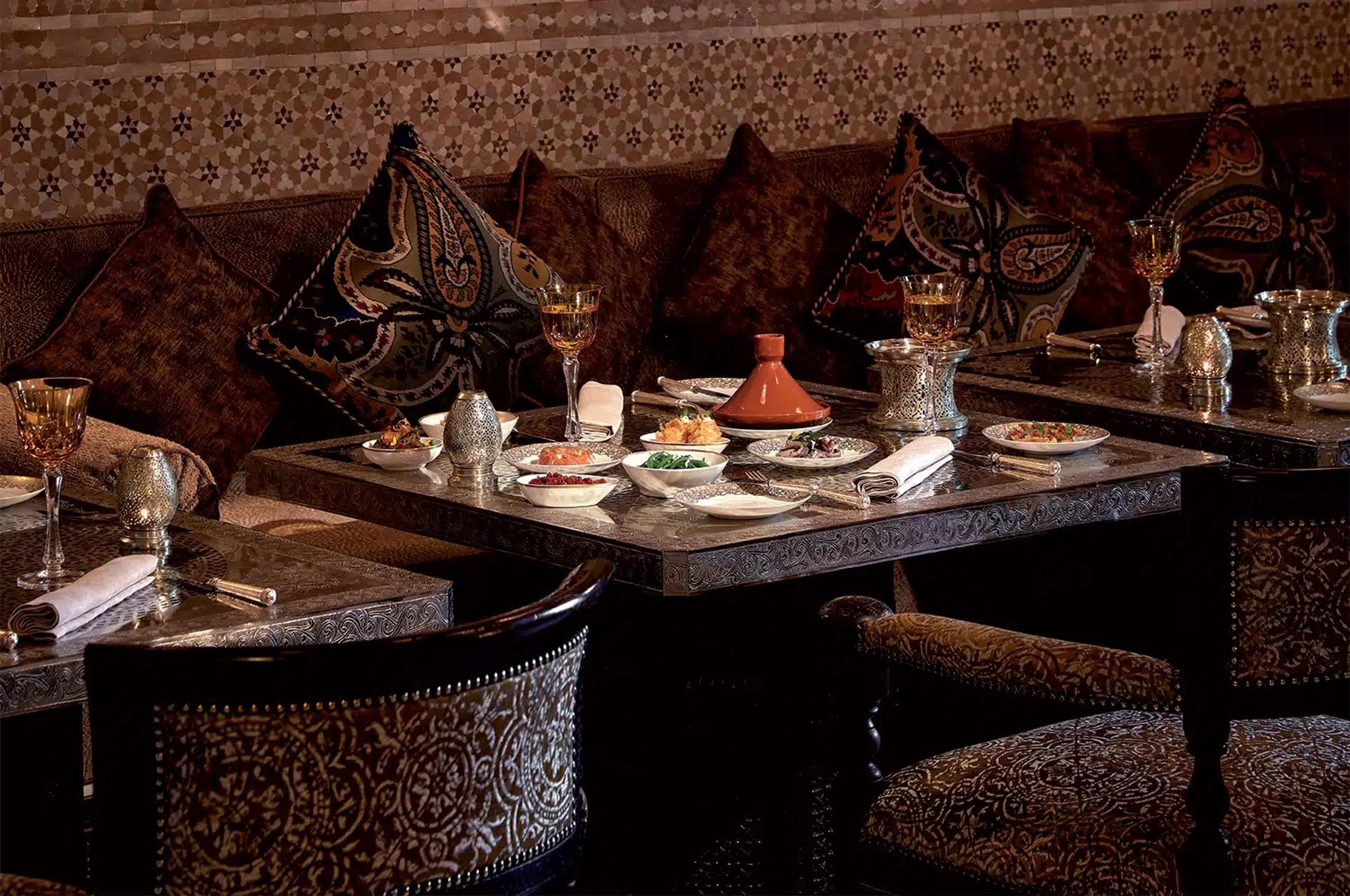 A carved, dimly lit Moroccan dining room with a table set beside a deep banquette under brass lanterns