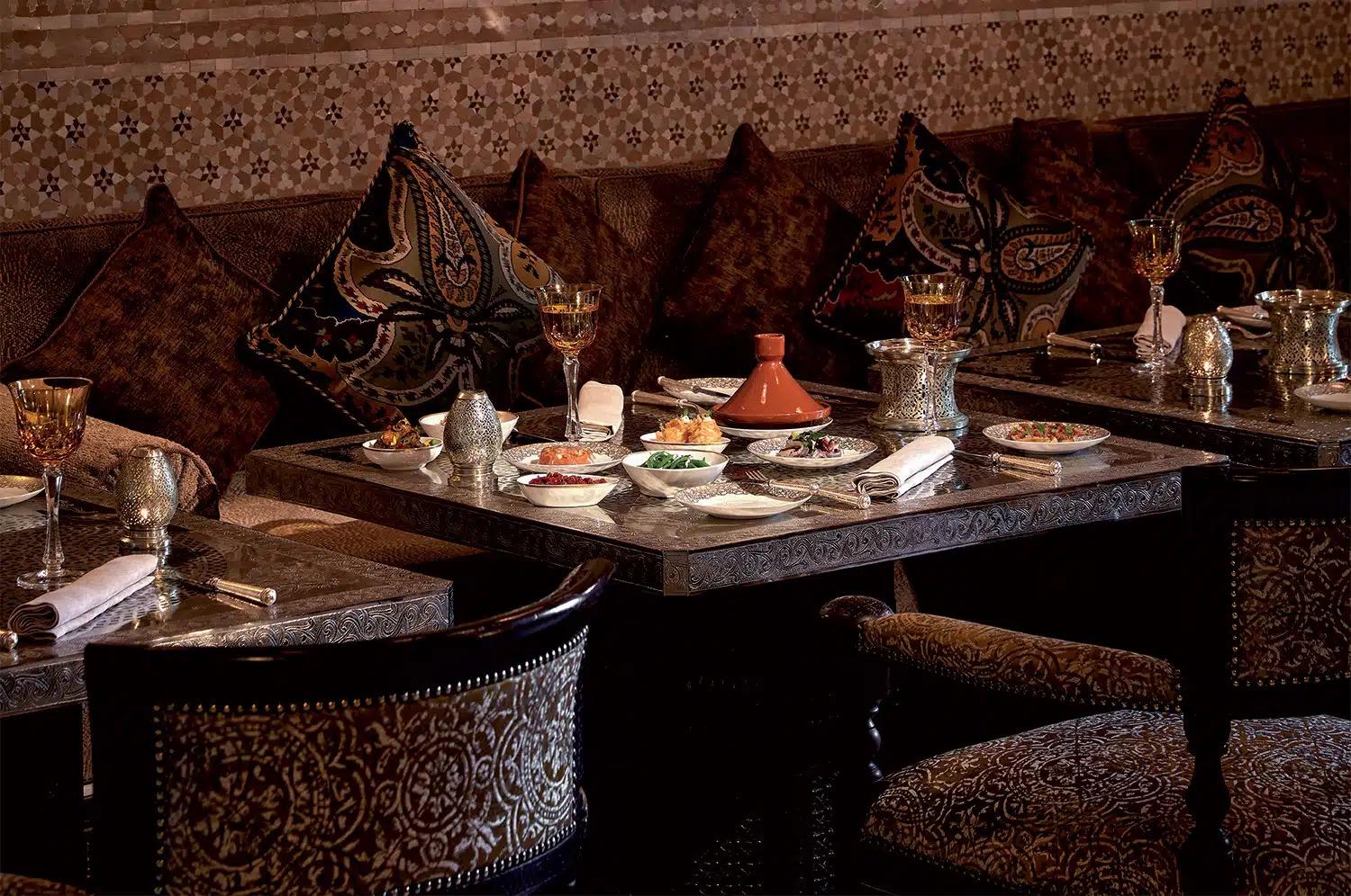 A carved, dimly lit Moroccan dining room with a table set beside a deep banquette under brass lanterns