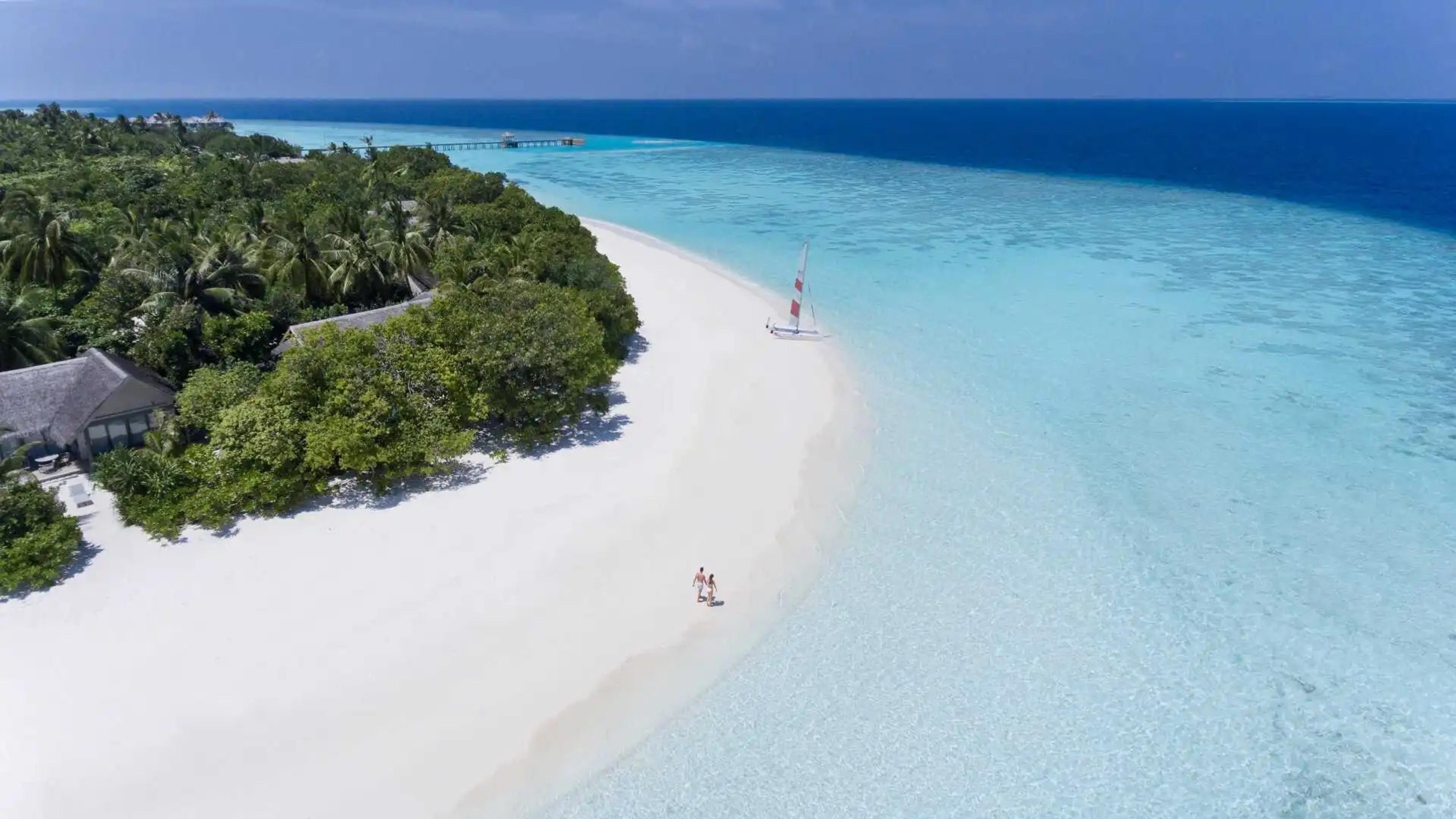 a stretch of white sand beach beside a shallow lagoon, with two people walking far down the shore and a small sailing catamaran pulled up on the sand
