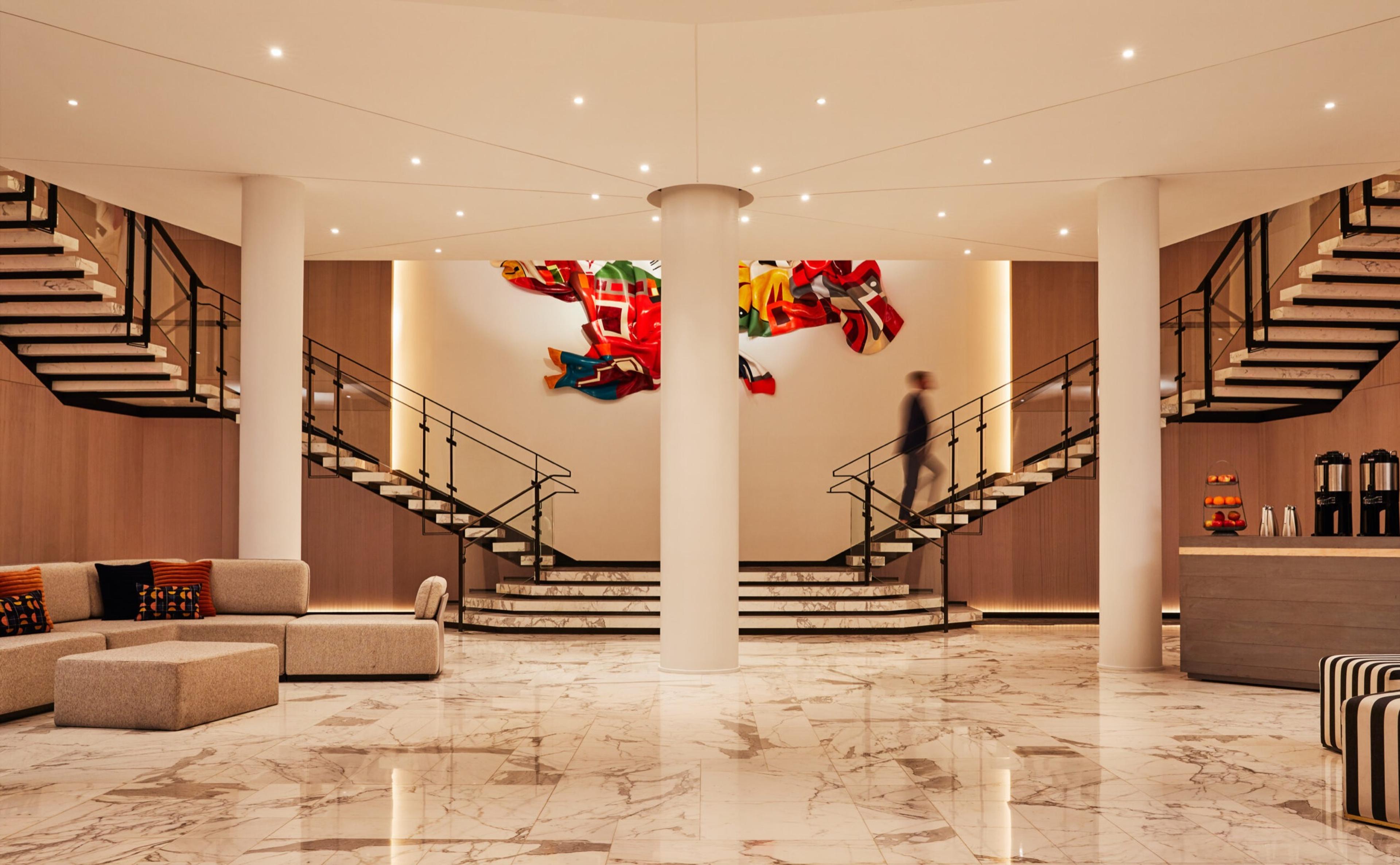 A hotel lobby with marble floors, two curved staircases, and a large abstract artwork on the wall, with one person walking across the space in the distance.