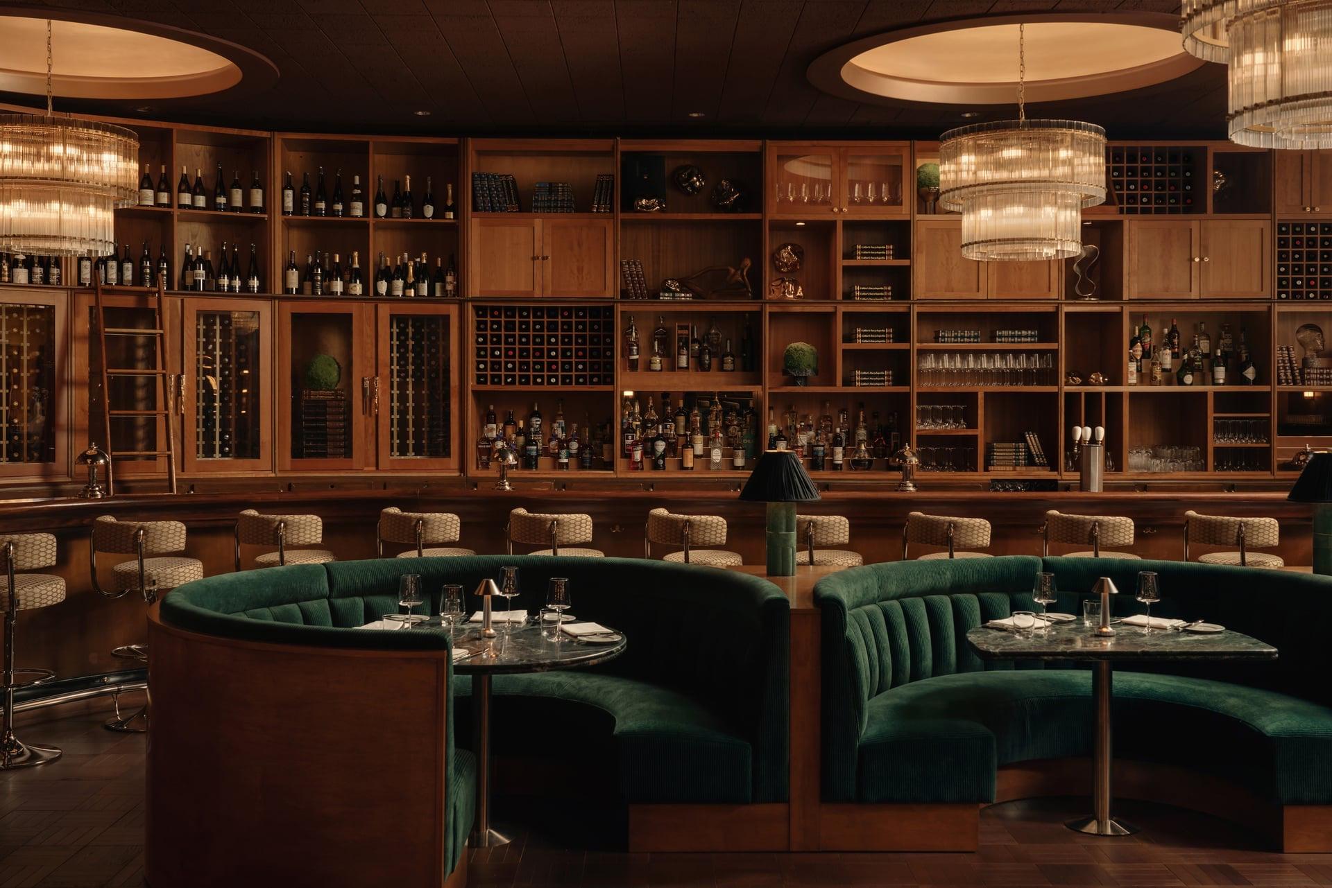 A dark wood bar room with green leather booths, chandeliers and shelves of bottles