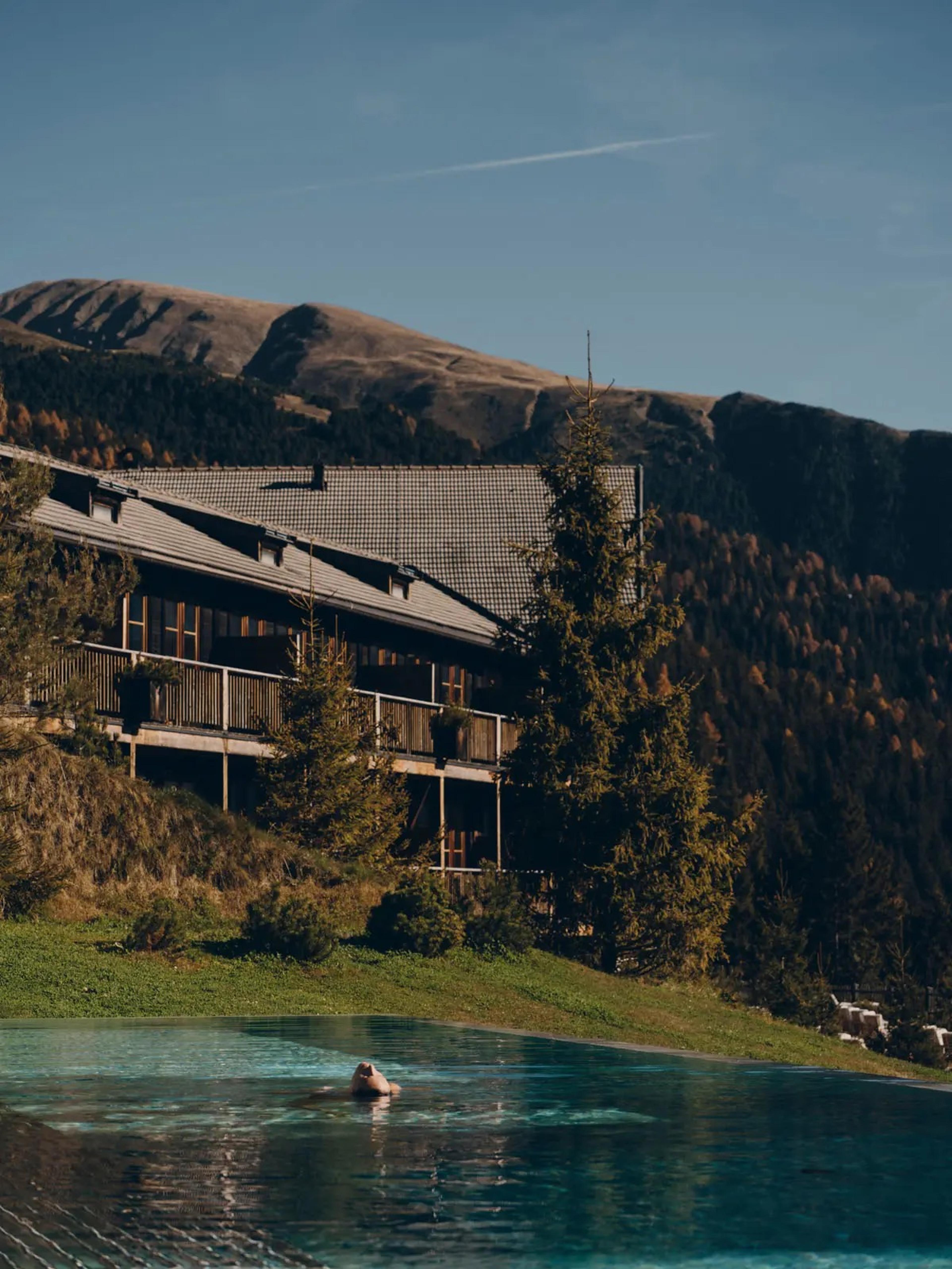 A long low timber hotel building sits above an infinity pool on a mountainside, with a lone swimmer and peaks beyond.