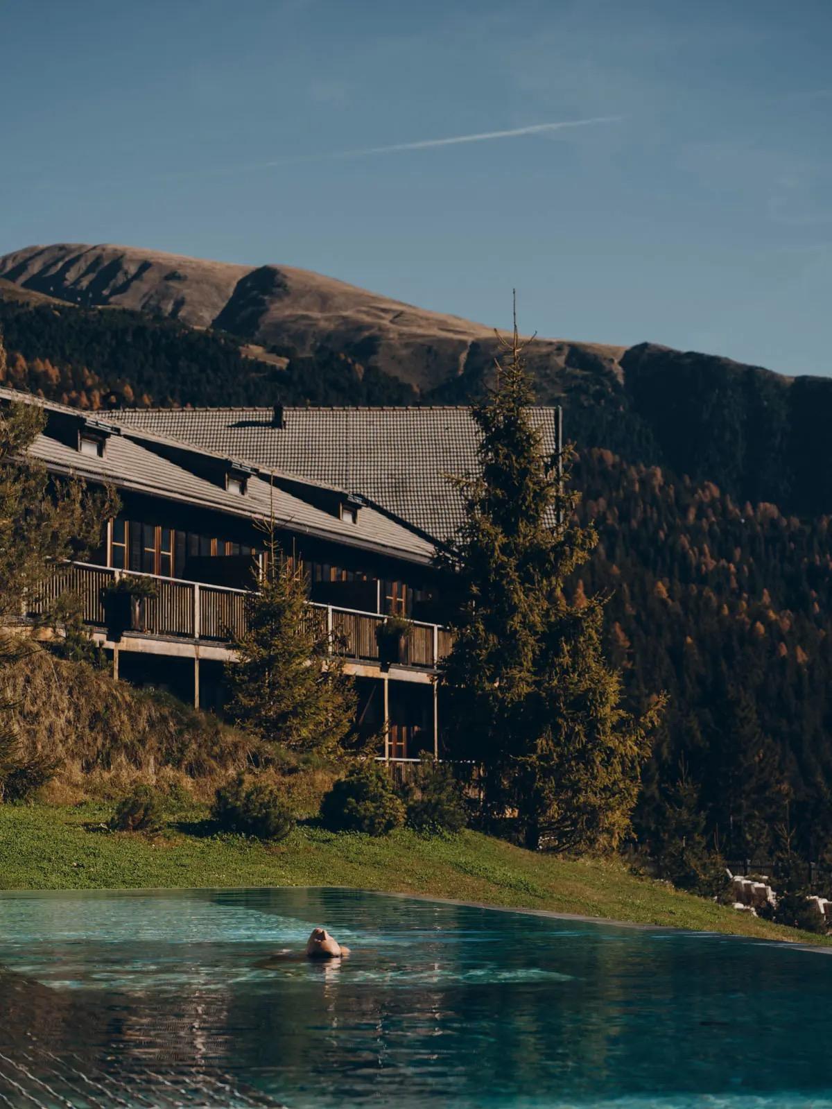 A long low timber hotel building sits above an infinity pool on a mountainside, with a lone swimmer and peaks beyond.