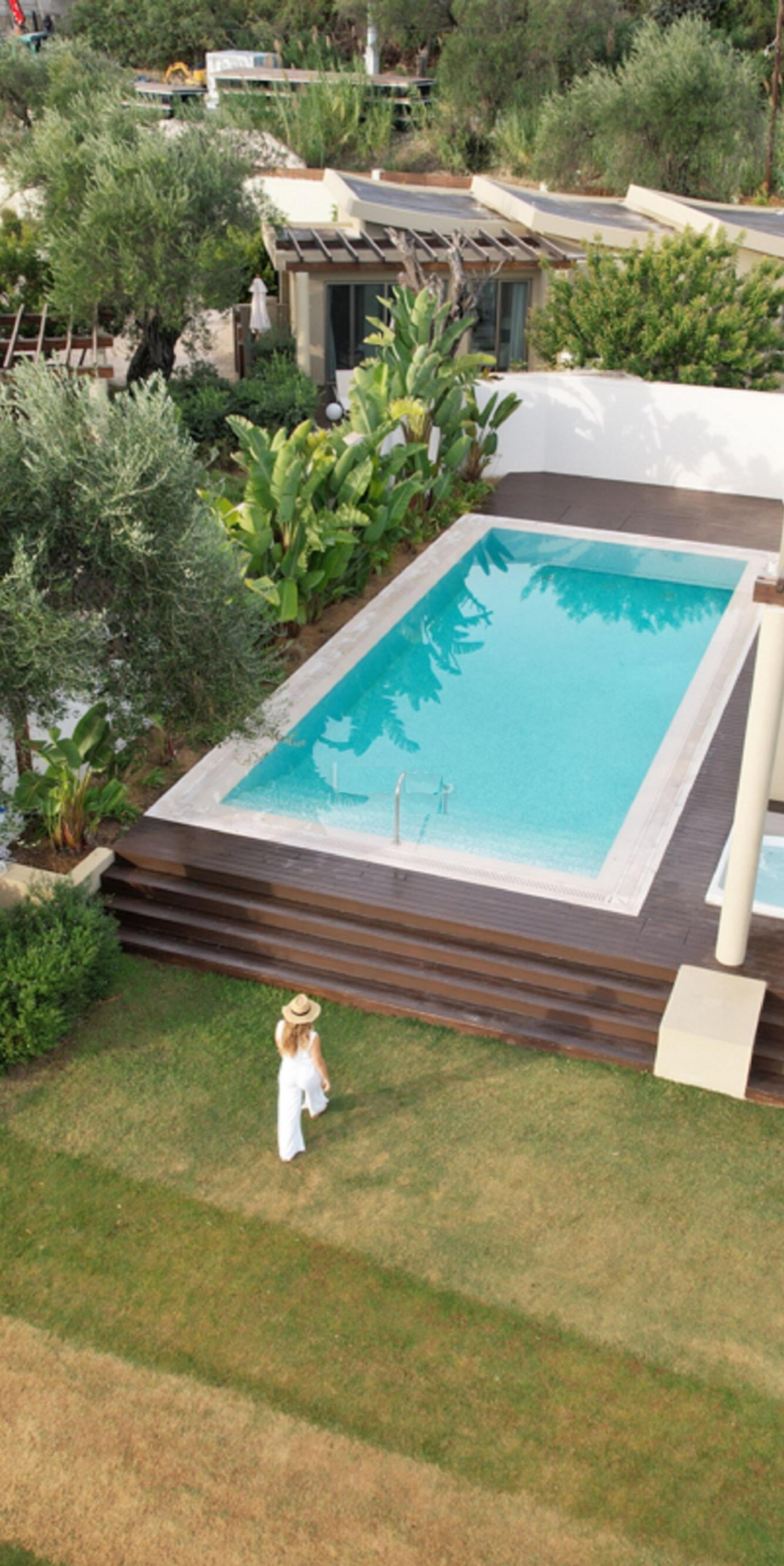 An aerial view of a rectangular plunge pool on a wooden deck beside a villa, with a person in a white outfit and sunhat walking away across a lawn in the foreground.
