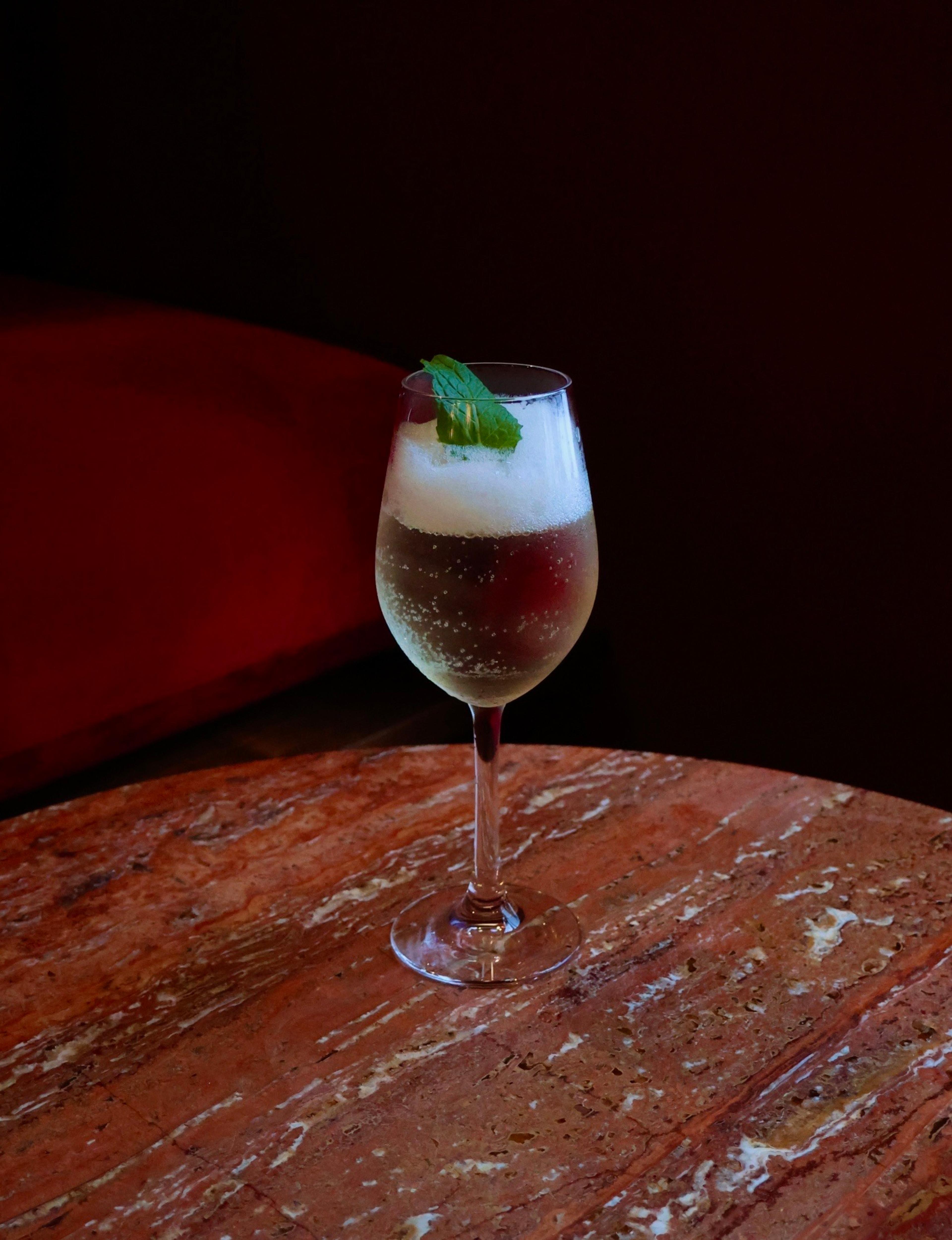 A champagne coupe garnished with mint on a marble table, with a red banquette behind it.