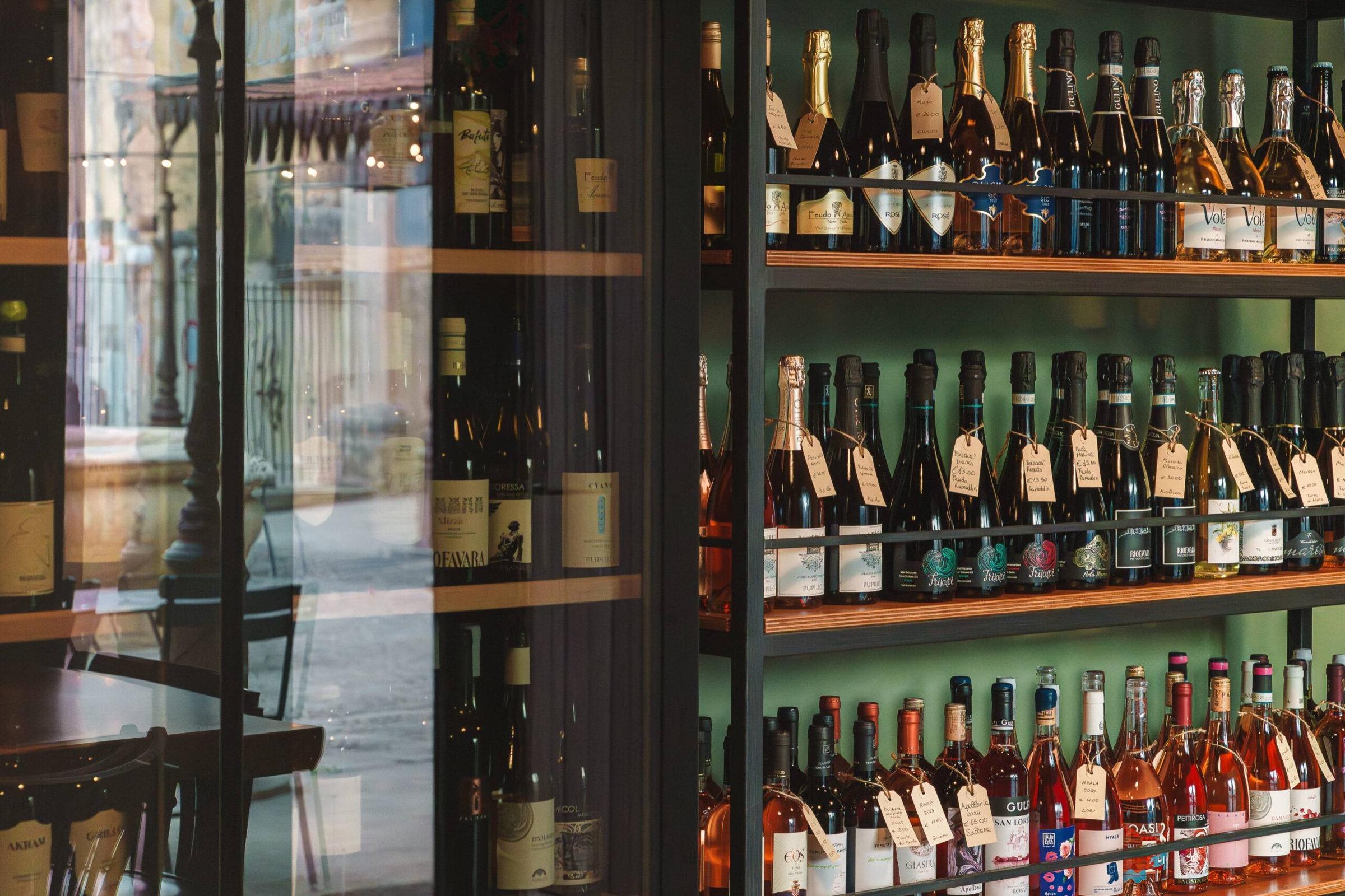 Shelves of wine and prosecco bottles with handwritten price tags behind a glass cabinet, reflecting a cobbled street and outdoor tables.