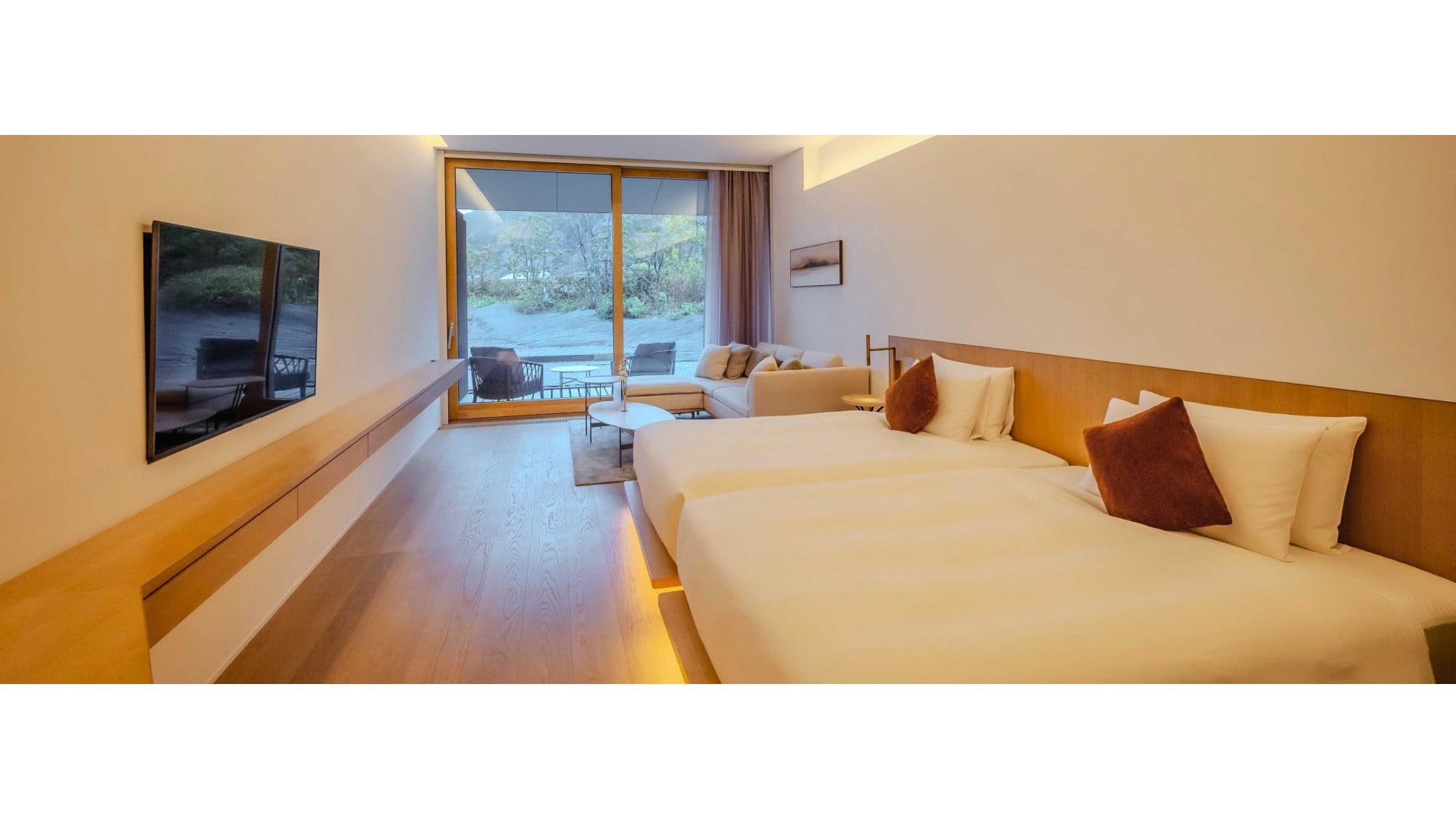 A hotel bedroom with two beds, a wall-mounted television, and a sliding glass door onto a wooded terrace