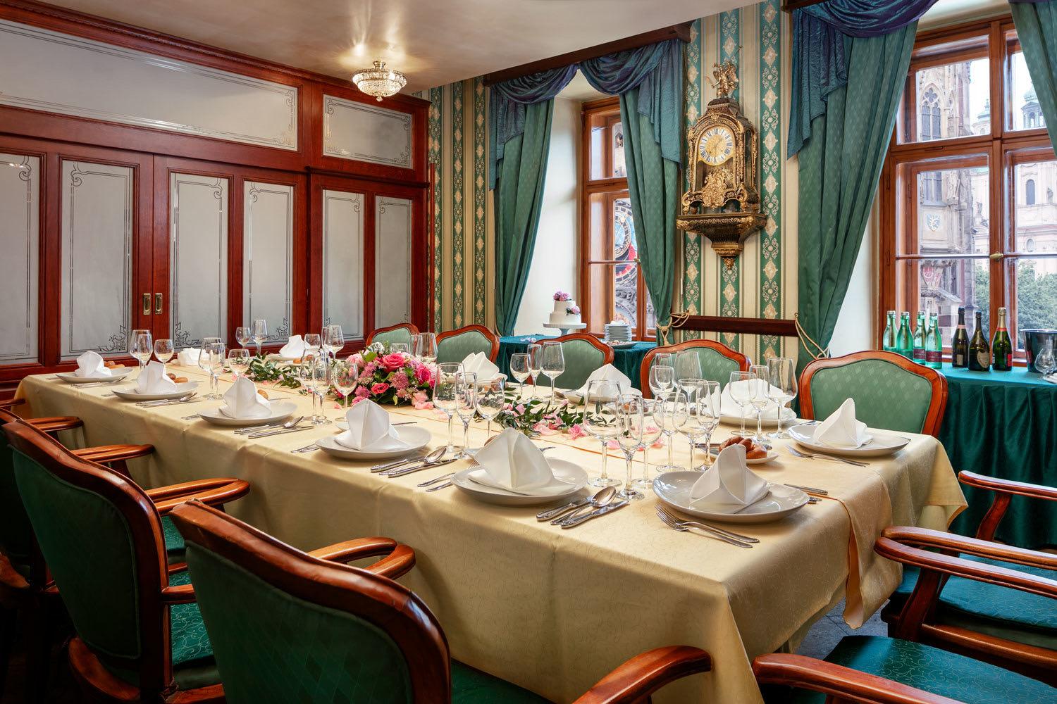 A long dining table set with white linen, wine glasses and flowers, in a room with green curtains and a wall clock.