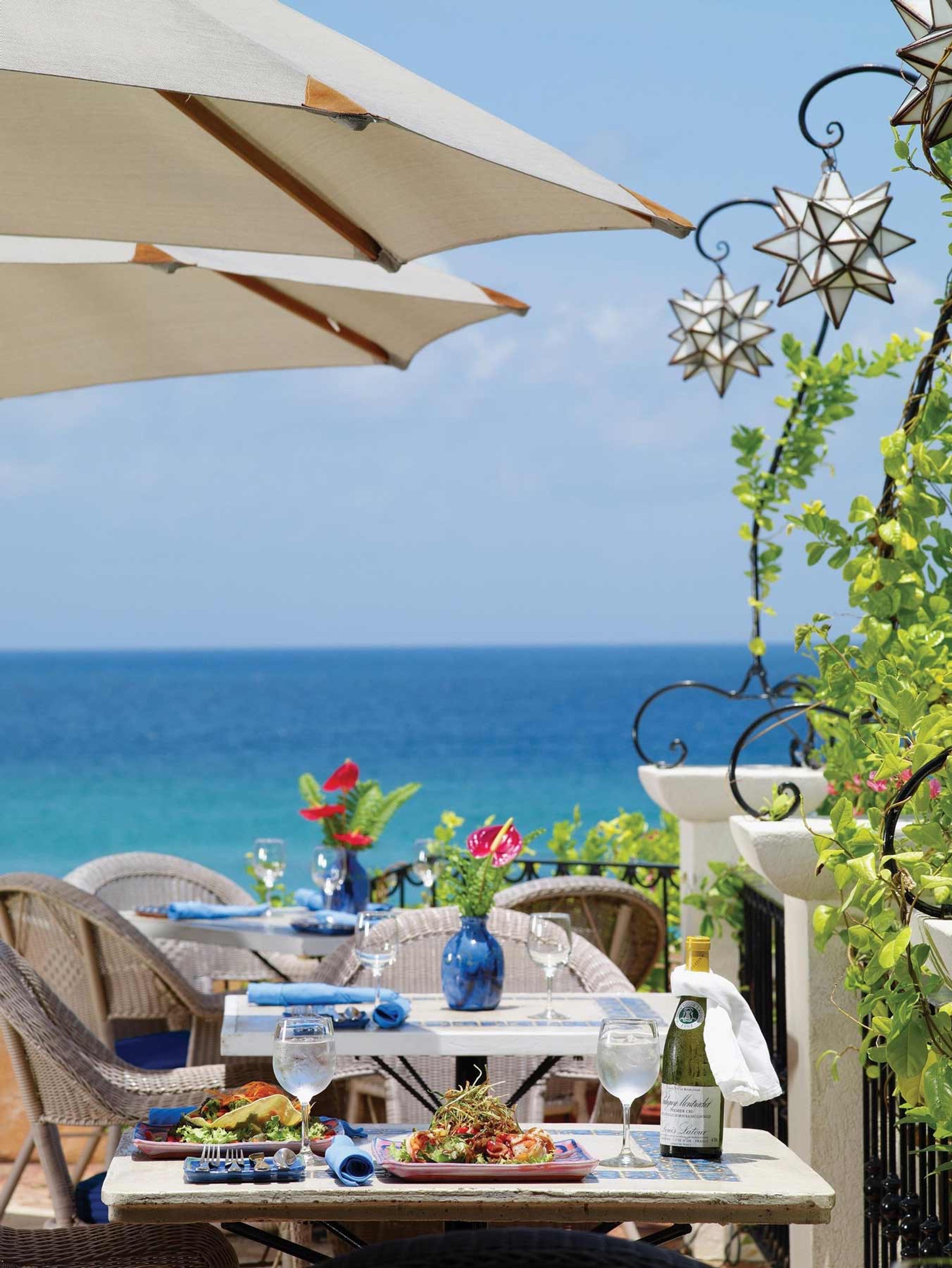 An oceanfront table set with salads, wine glasses and a bottle, shaded by umbrellas with star shaped lanterns hanging nearby.