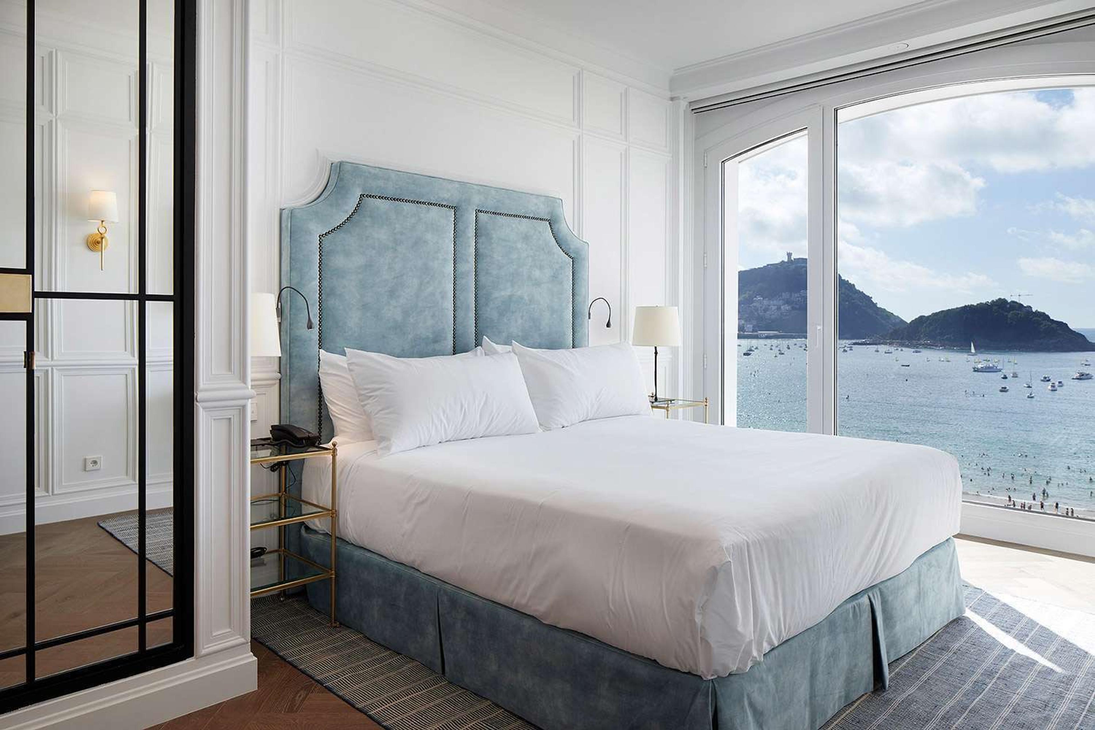 A bedroom with a pale blue upholstered headboard and an arched window open to a view of the bay and its boats.