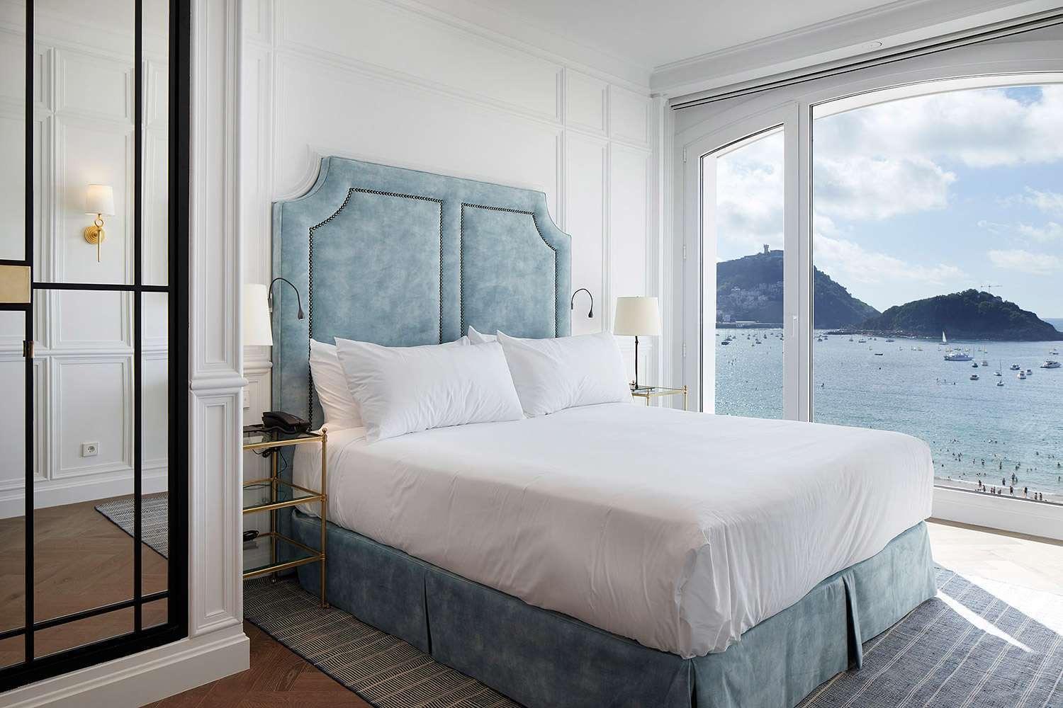 A bedroom with a pale blue upholstered headboard and an arched window open to a view of the bay and its boats.