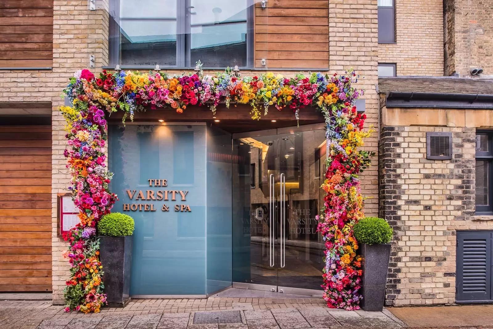 the hotel's glass front door framed by an arch of colourful artificial flowers, with the name The Varsity Hotel and Spa on the wall beside it