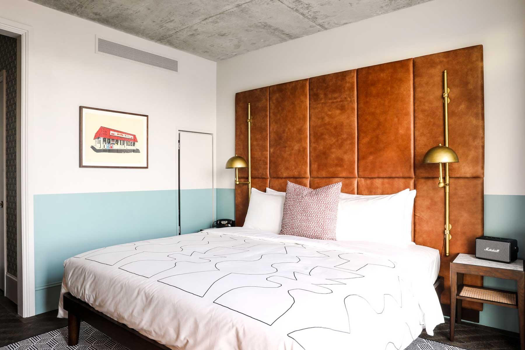 A hotel bed with a tan leather headboard under an exposed concrete ceiling, with a framed print of a red brick building on the pale blue wall beside it.