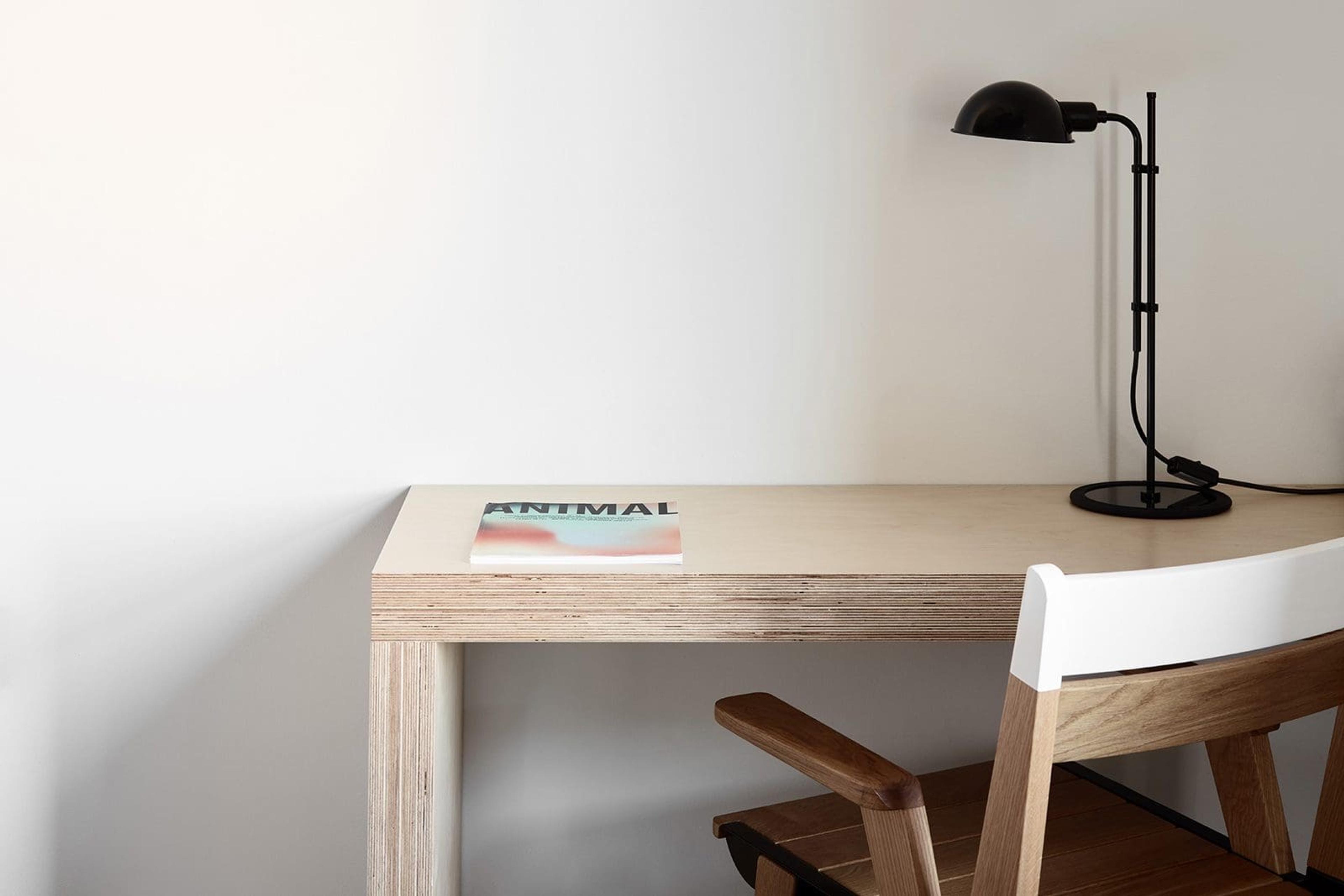 a plywood desk against a white wall holding a magazine and a black articulated desk lamp, with a wood and leather chair tucked underneath