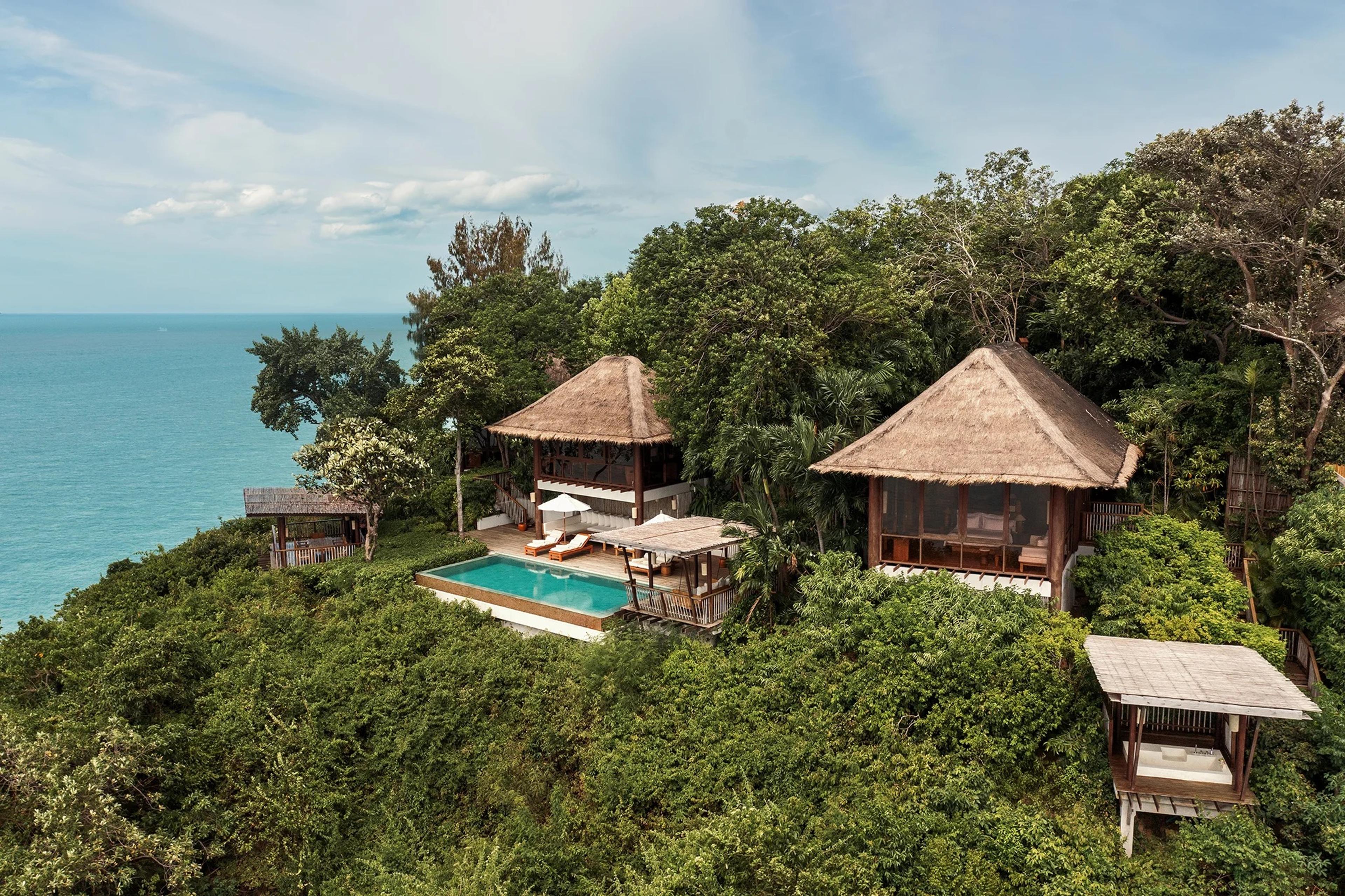 An aerial view of a hillside villa with two thatched roofs, a private pool and an open-sided lounge, surrounded by dense trees above the sea.