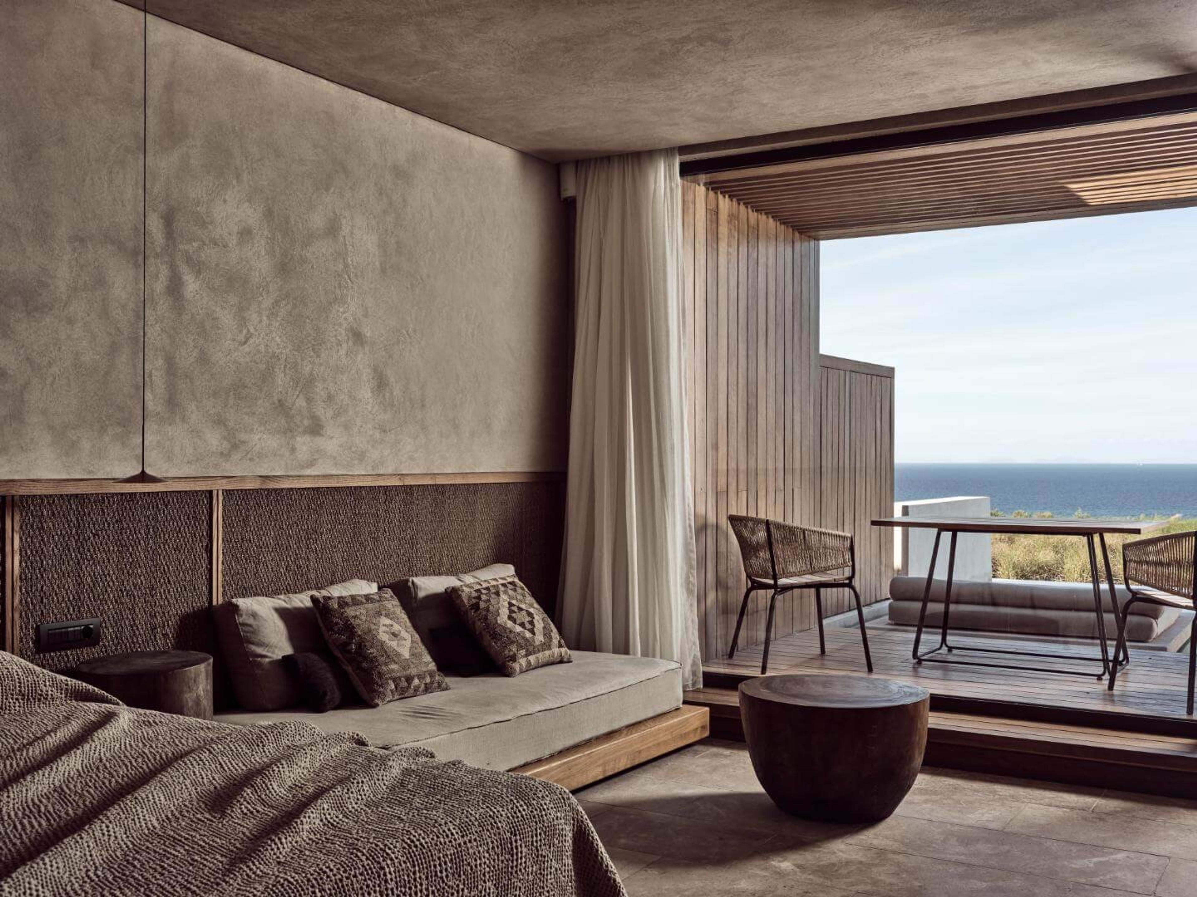 A bedroom with a grey throw and patterned cushions opens directly onto a wooden terrace with chairs and the sea beyond.
