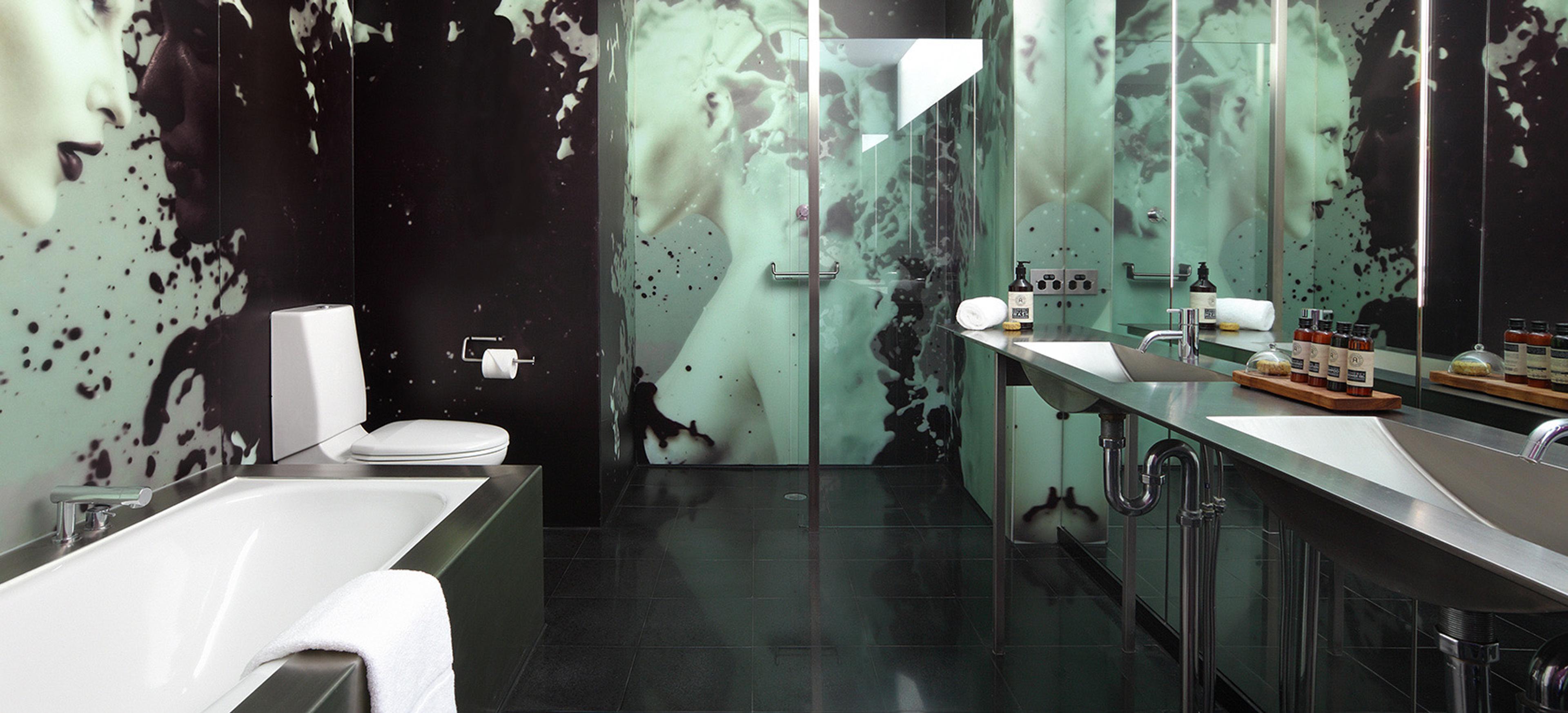 A bathroom with a black and white mural of a face across the walls, a built in tub, and a glass shower beside twin sinks.
