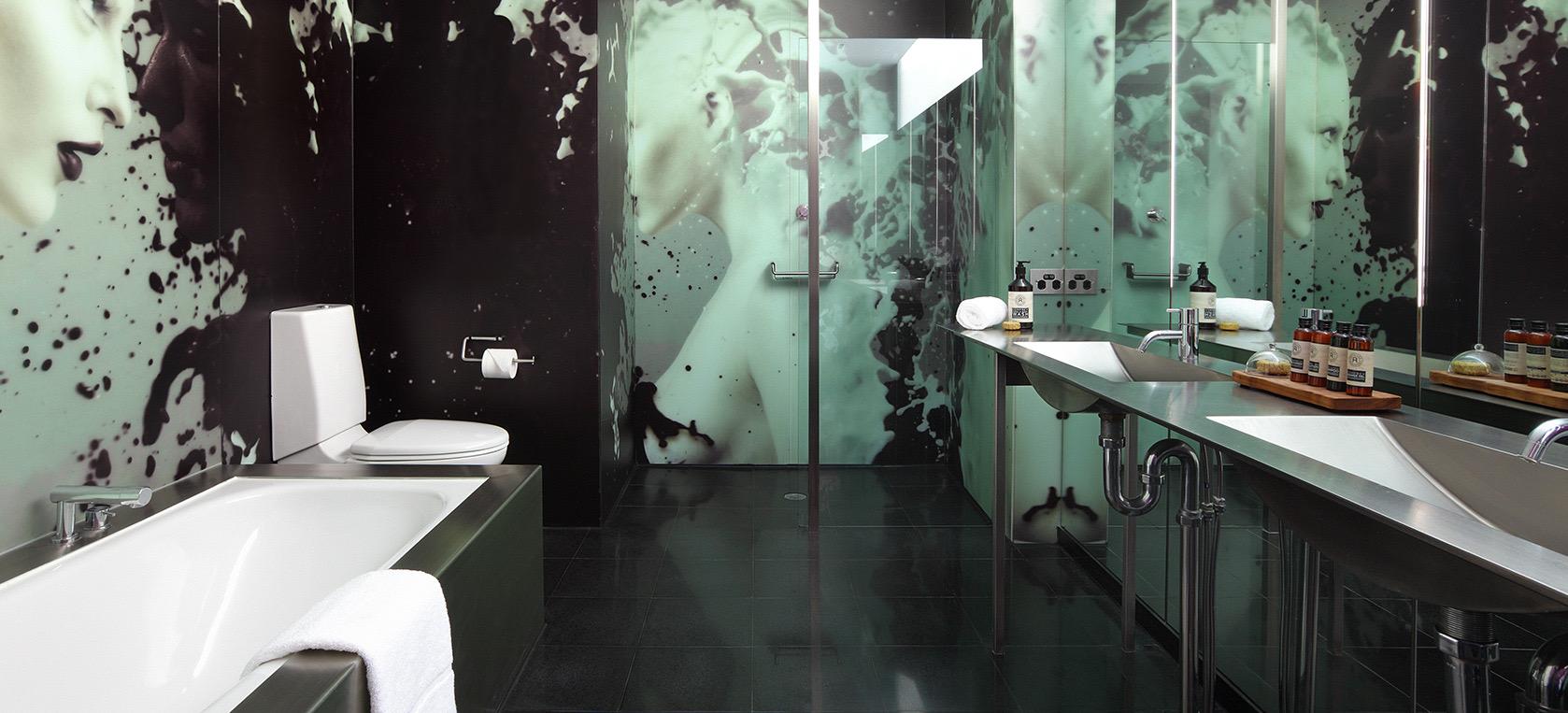 A bathroom with a black and white mural of a face across the walls, a built in tub, and a glass shower beside twin sinks.