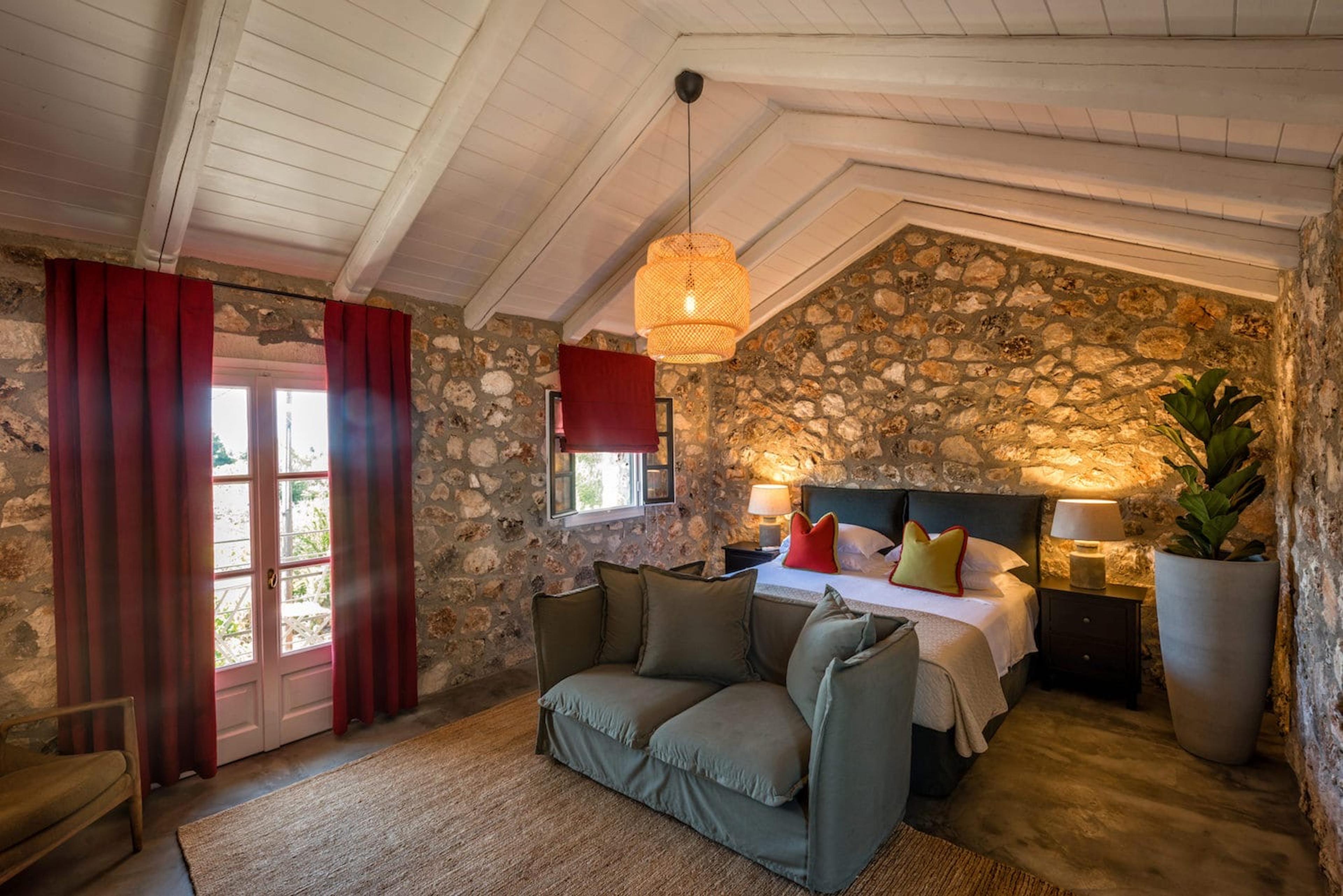 An attic bedroom with a beamed ceiling, a stone wall, a sofa at the foot of the bed, and red curtains open onto French doors with daylight beyond.